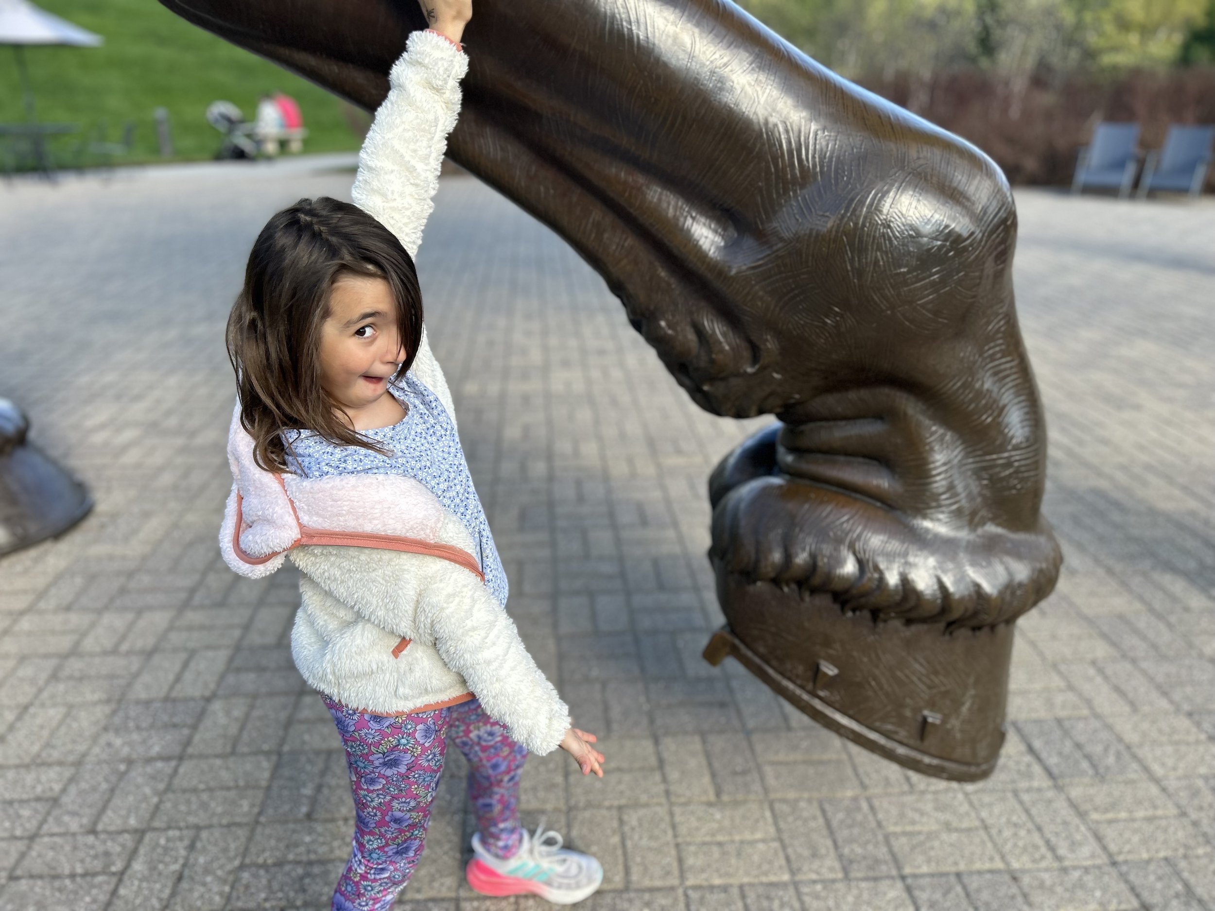 A young girl with long brown hair wearing a white fuzzy jacket, patterned blue and pink pants, and pink shoes standing outdoors and touching a large bronze statue of a dog; the background shows a park with grass, trees, benches, and people.