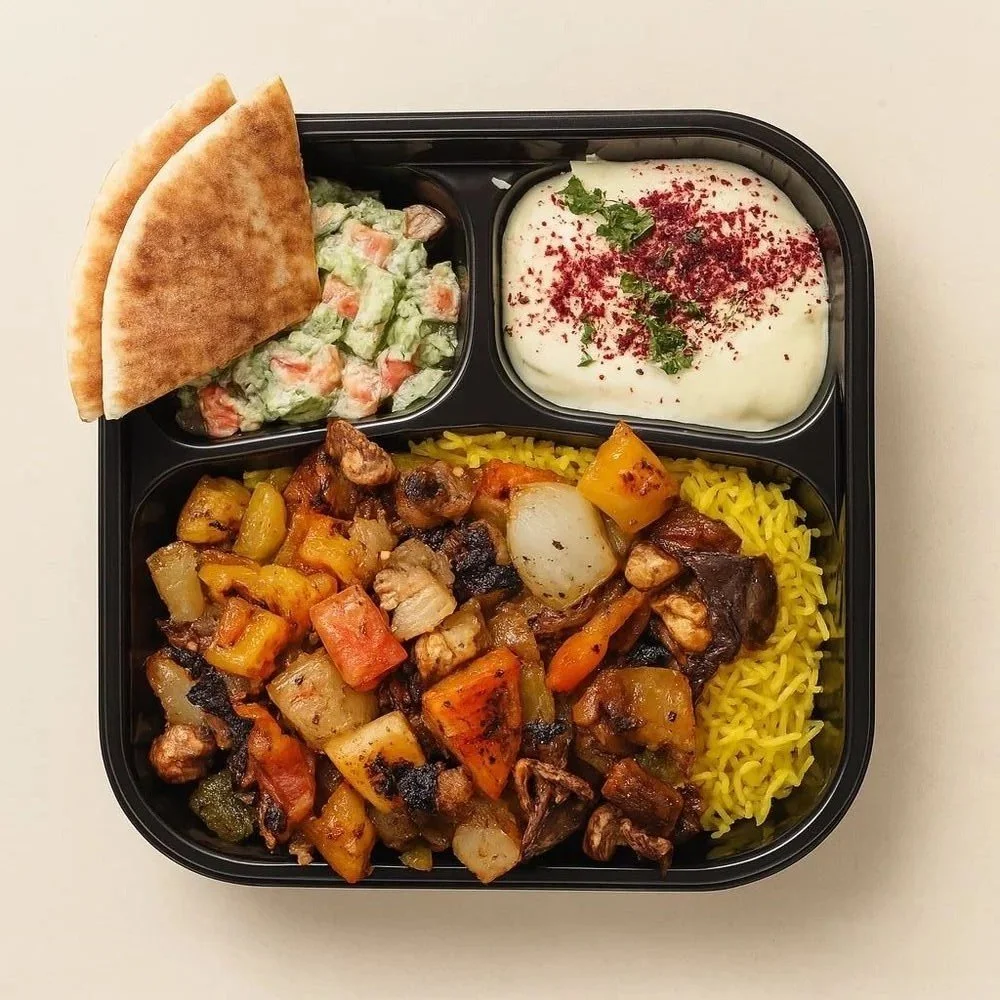 A black meal prep container with yellow rice, cooked vegetables and chicken, a side of cucumber and tomato salad, a serving of yogurt with red powder and mint, and two pita bread halves.