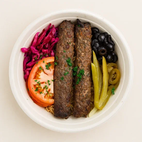 A bowl of Mediterranean food with two grilled kebabs, tomato slice with herbs, pink pickled cabbage, black olives, green pepper slices, and onion slices on a white background.