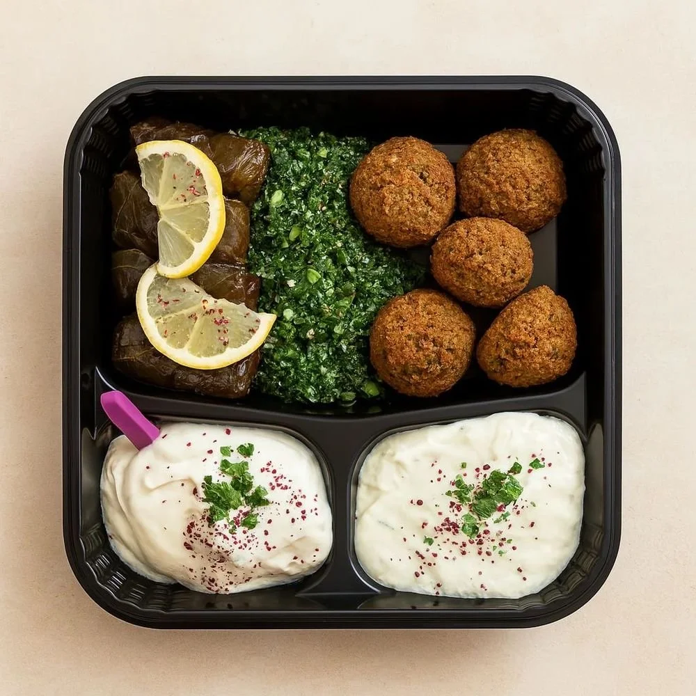 A black compartmentalized dish with stuffed grape leaves garnished with lemon slices, a portion of chopped green herbs, five falafel balls, and two servings of tzatziki sauce garnished with herbs and paprika.