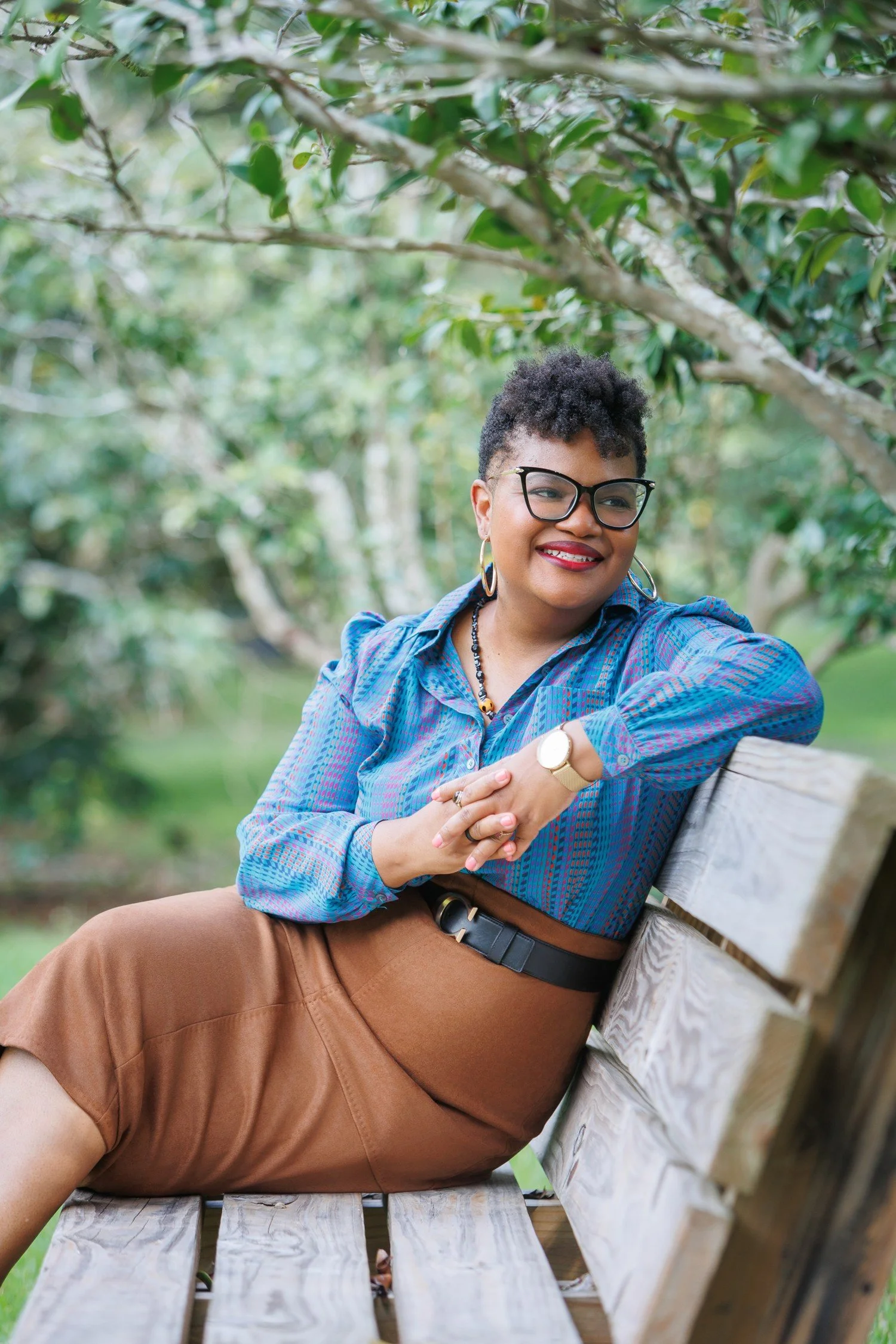 A woman with curly hair wearing glasses, a blue patterned blouse, and brown skirt, sitting on a park bench outdoors, smiling with greenery in the background.