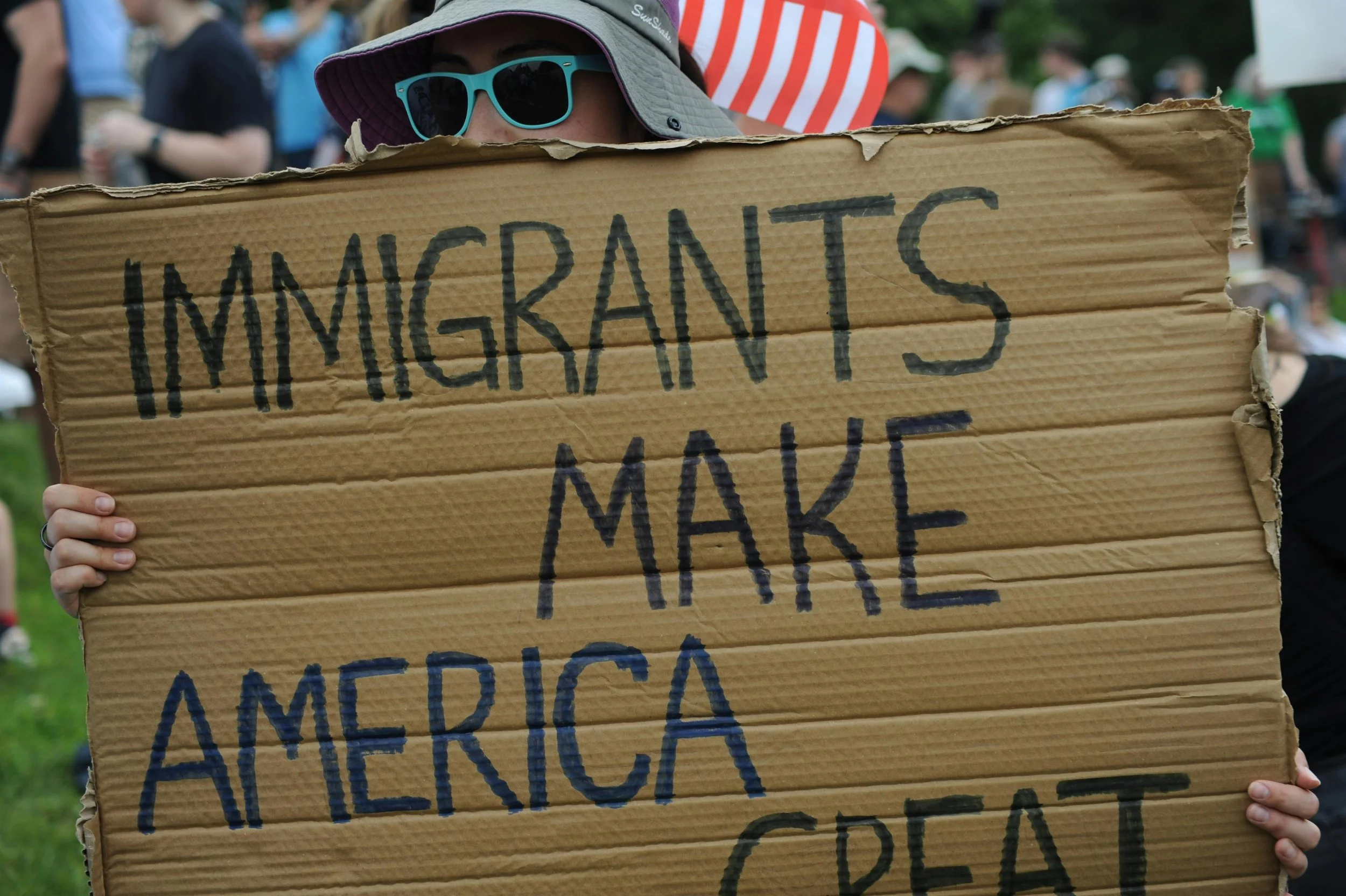 Person in a wide-brimmed hat and sunglasses holding a cardboard sign with handwritten text that reads "IMMIGRANTS MAKE AMERICA" at an outdoor event or protest.