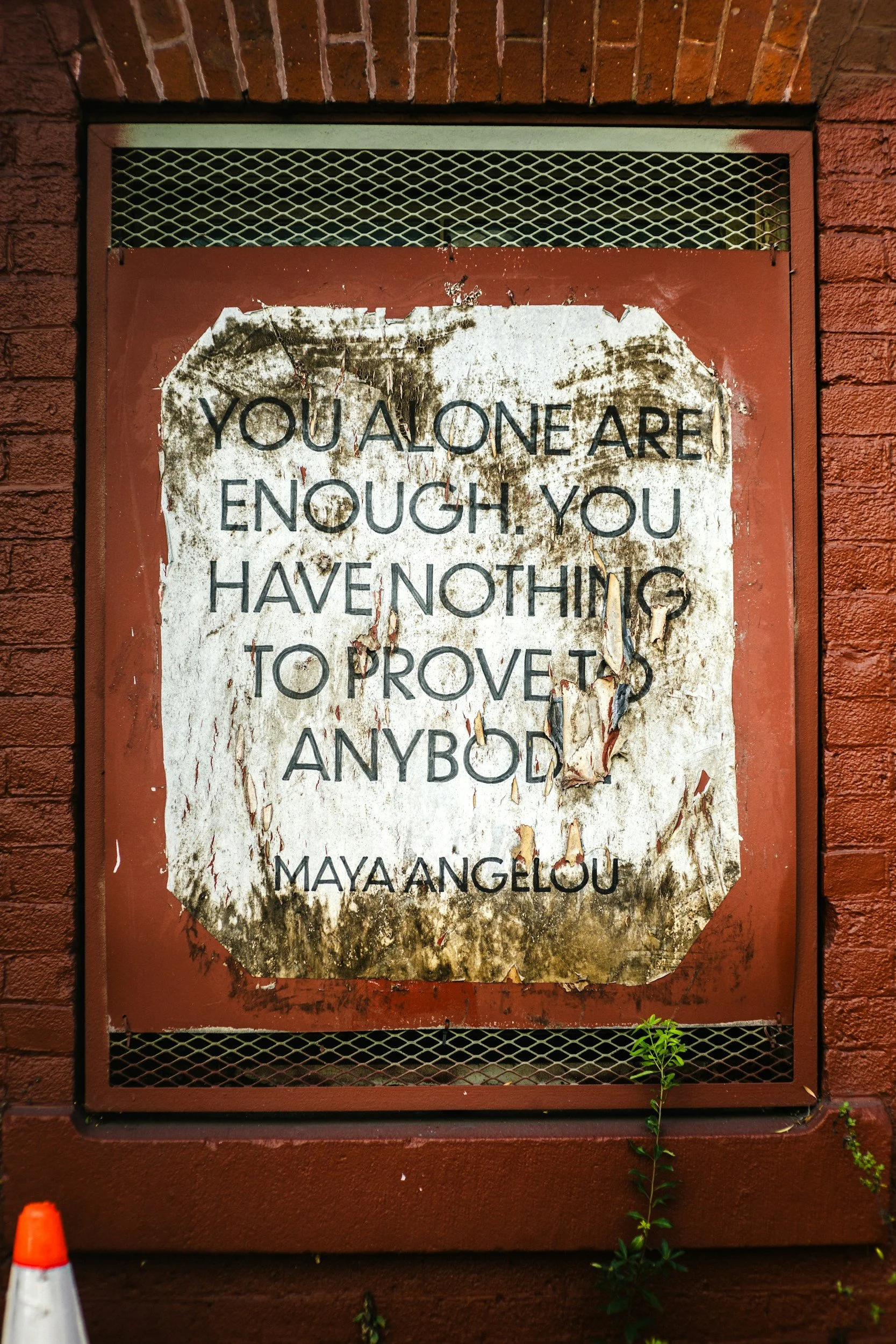 A worn, weathered sign on a red brick wall with incomplete and peeling text that reads, 'YOU ALONE ARE ENOUGH. YOU HAVE NOTHING TO PROVE TO ANYBODY.'