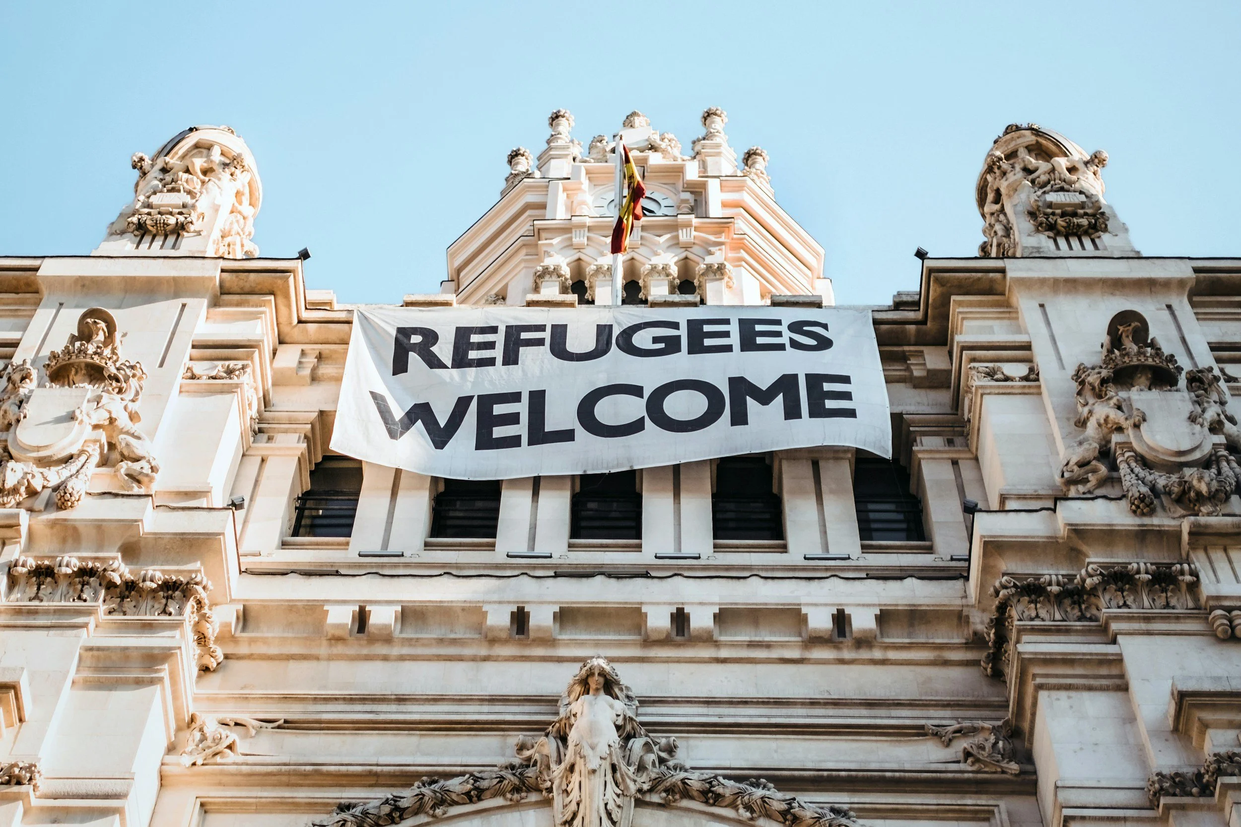 Historic building with ornate decorative sculptures and a large banner that reads 'Refugees Welcome' hanging on its facade.
