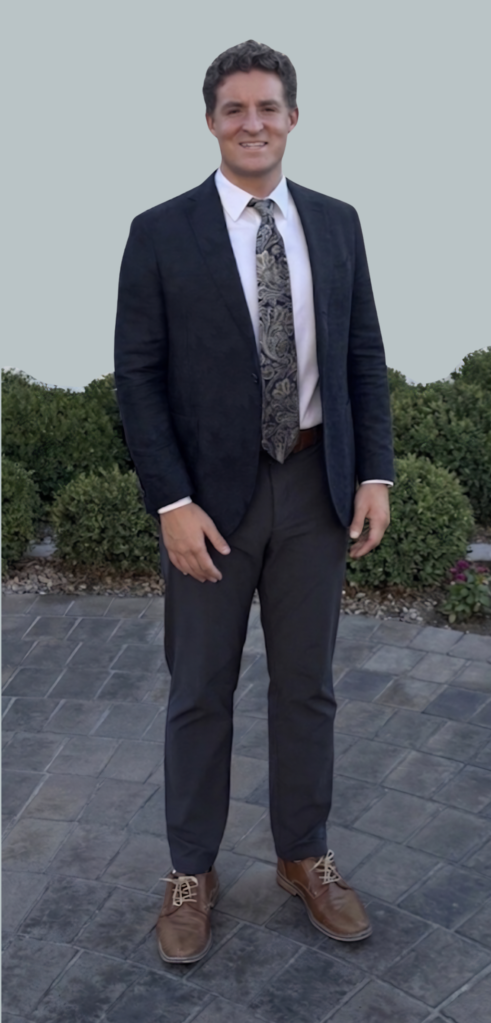 A man in a dark suit with a white shirt and patterned tie, standing outdoors on a paved area with bushes and flowers in the background.