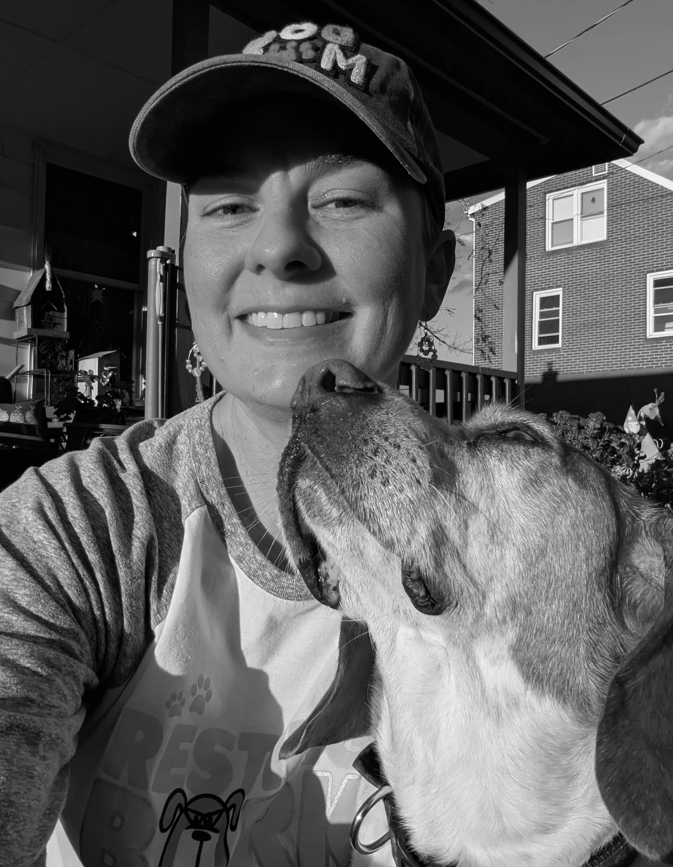 A woman with short hair wearing a baseball cap and earrings is taking a selfie outdoors with her dog, who is licking her face. In the background, there is a house with a brick exterior and a deck with falling leaves.
