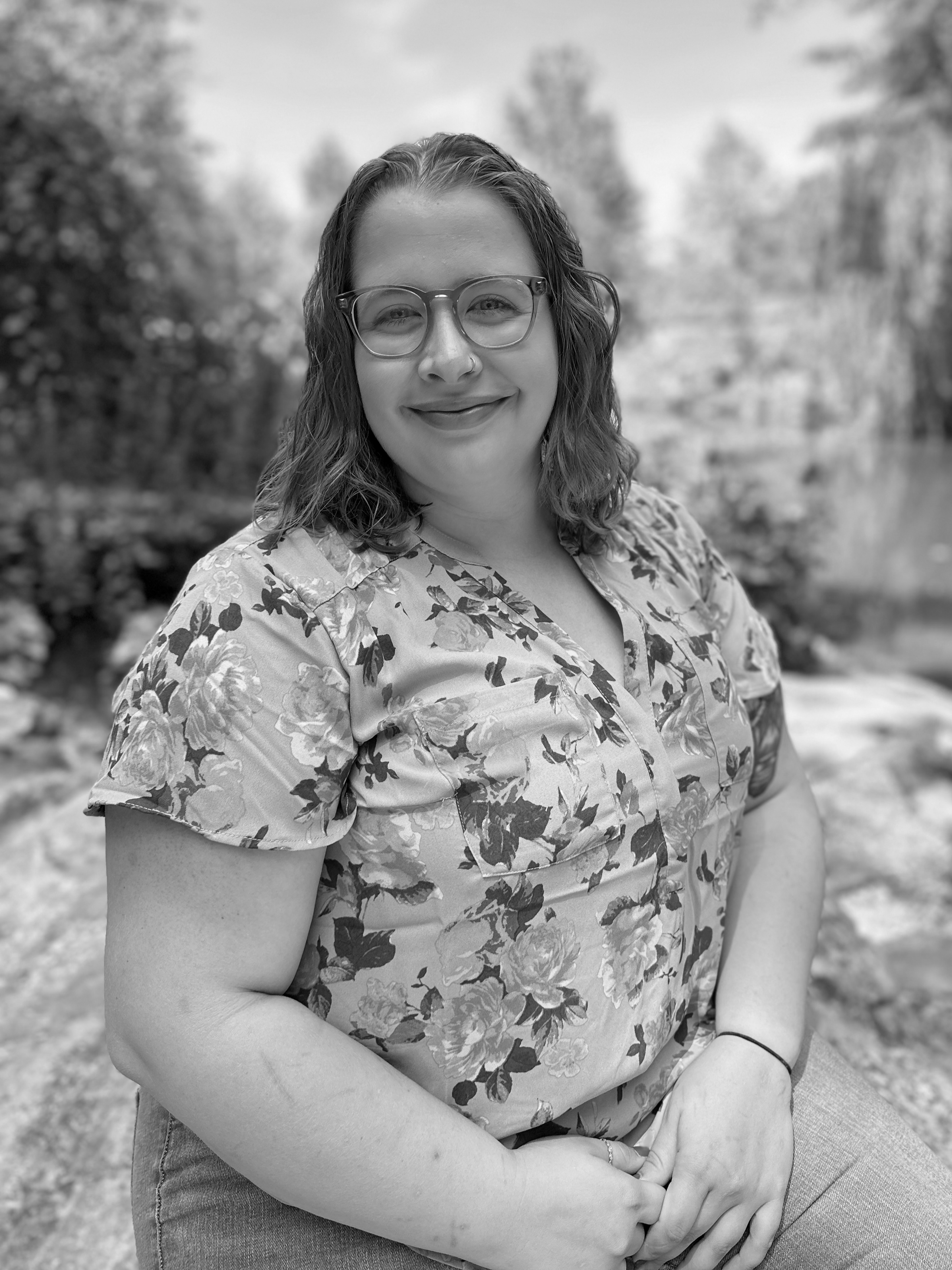 Black and white photo of a woman with glasses smiling outdoors, wearing a floral shirt, with trees and a body of water in the background.