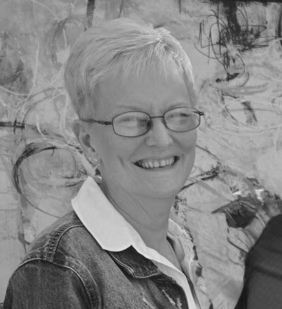 A smiling woman with short, light hair and glasses, wearing a denim jacket over a collared shirt.
