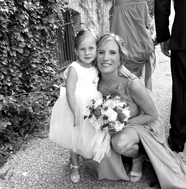 A woman and a young girl, both smiling, pose together outdoors. The woman is holding a bouquet of flowers, and the girl is wearing a light-colored dress. They are standing near a vine-covered fence with other people partially visible in the background.