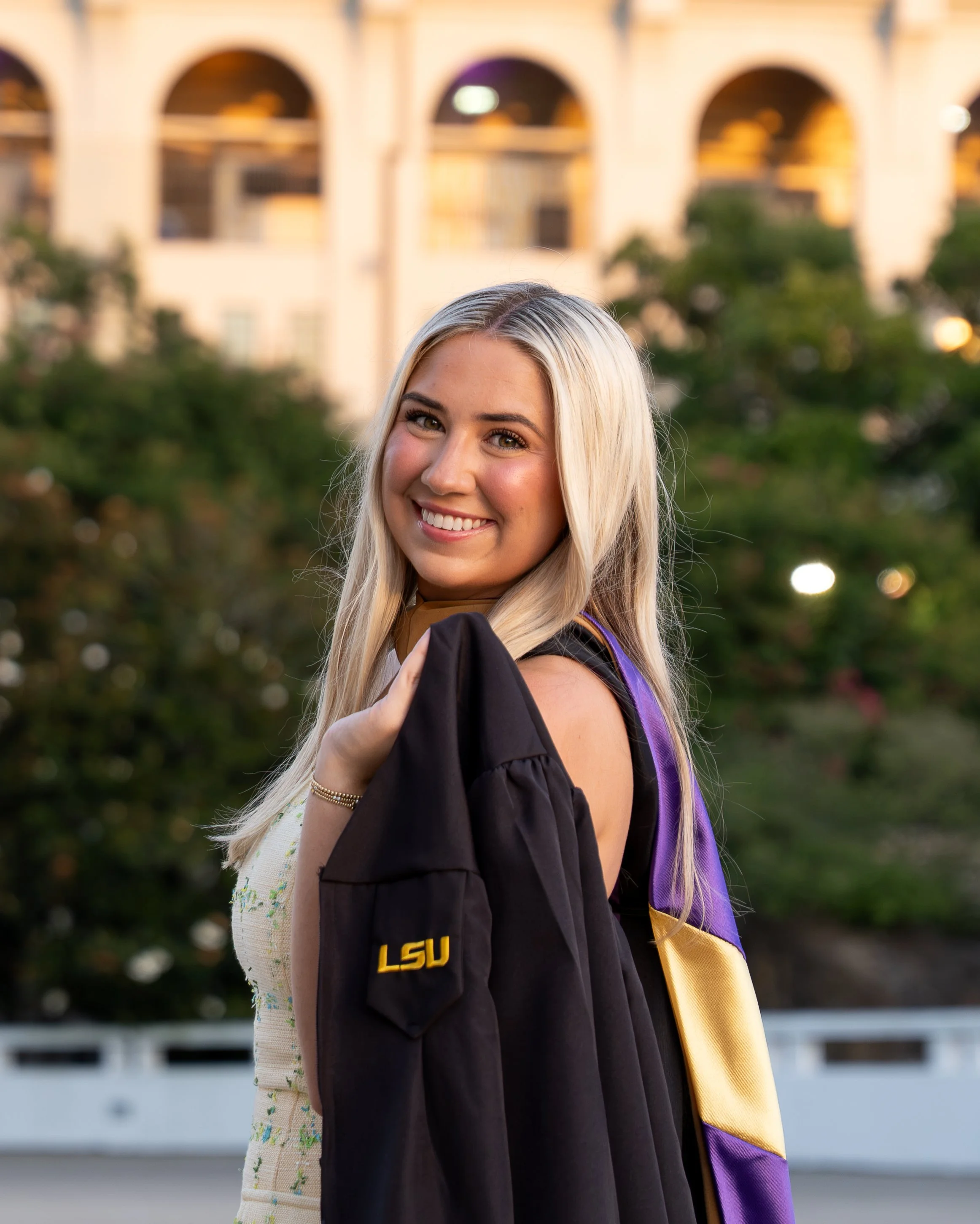 A young woman with long blonde hair, smiling, in her graduation cap and gown, holding her diploma, with a university building and trees in the background.