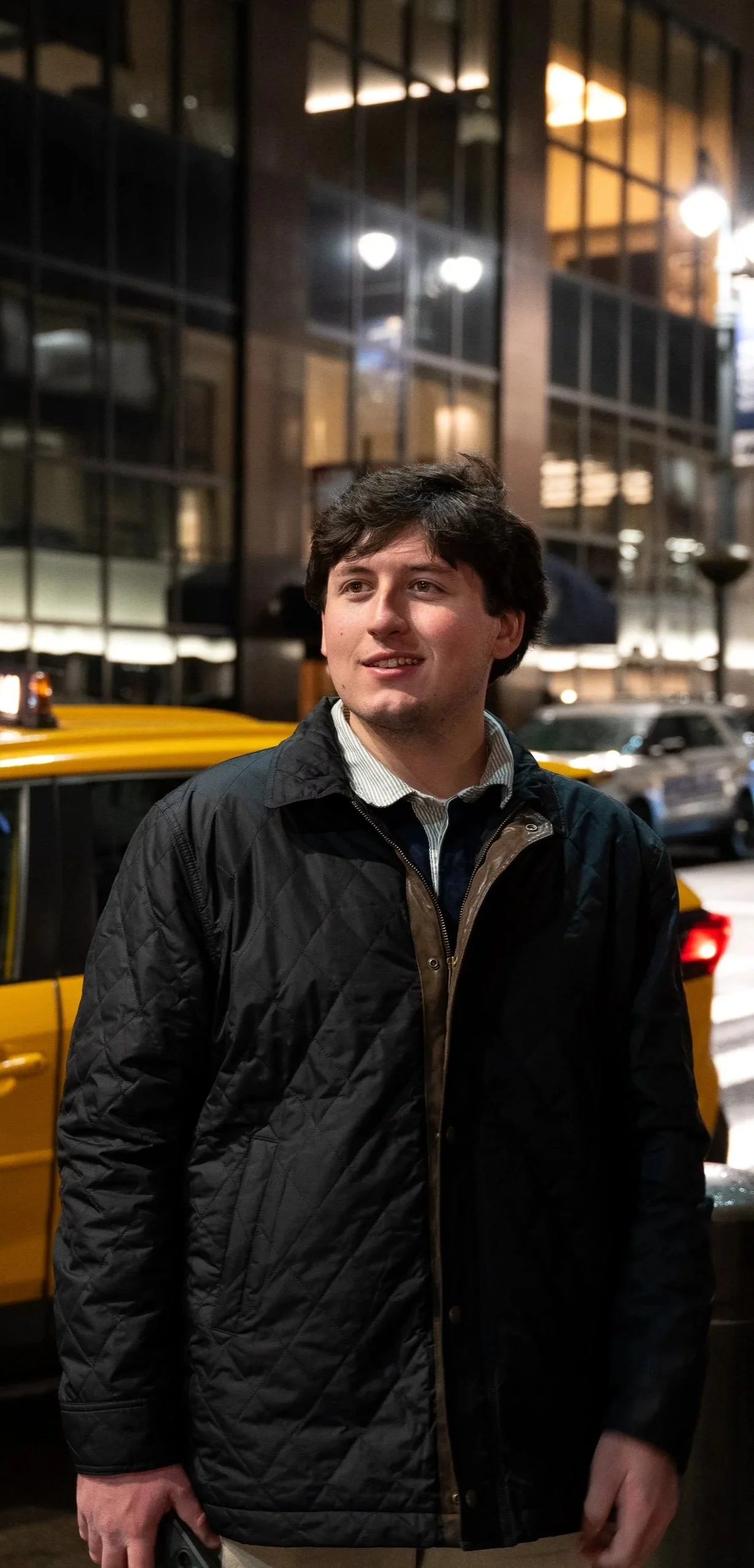 A young man with dark hair and fair skin standing on a city street at night, wearing a black quilted jacket over a collared shirt, with bright city lights and tall buildings in the background.