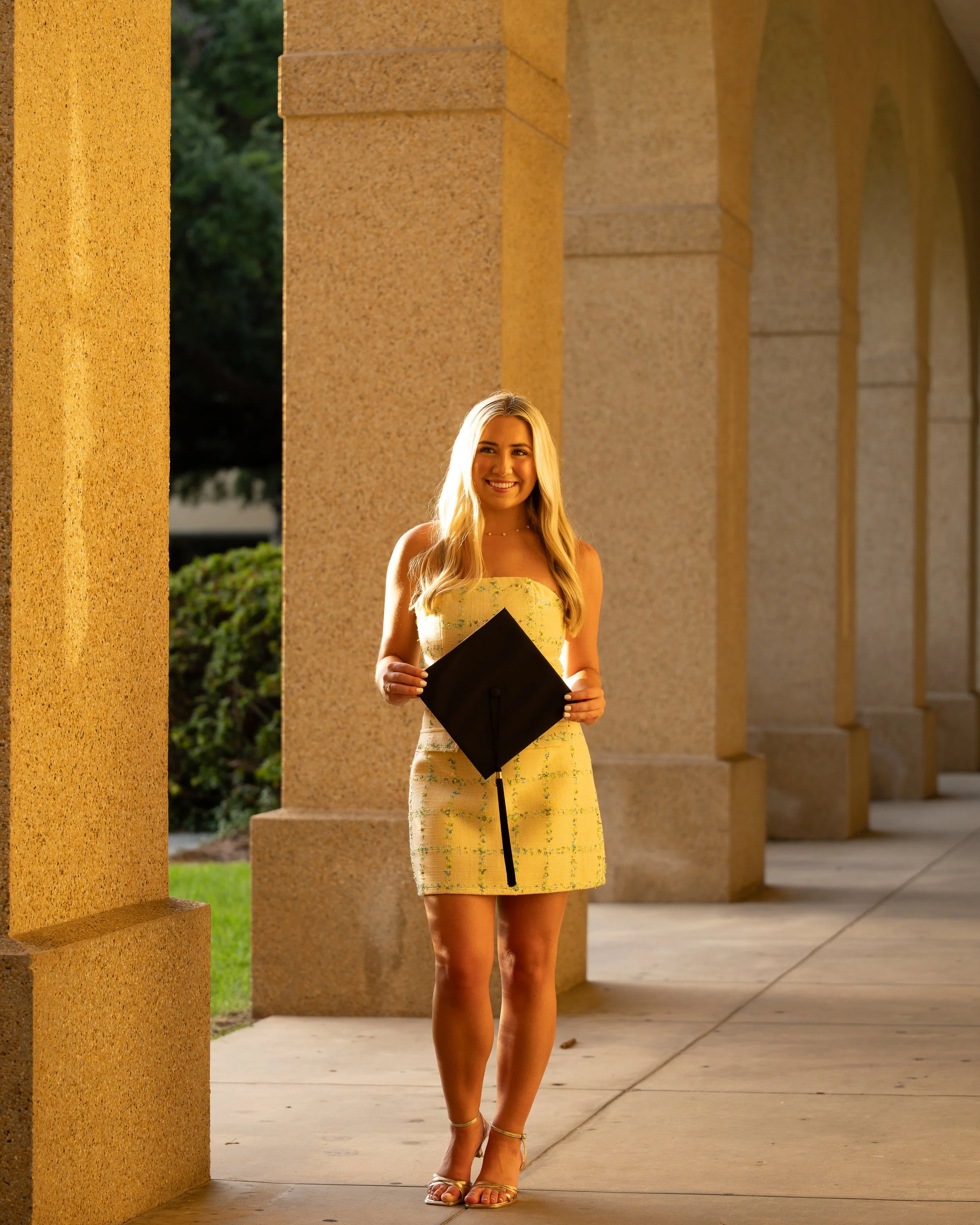 A young woman with long blonde hair, wearing a strapless yellow dress with green accents, stands outside under a series of beige stone arches. She is smiling and holding a black folder in her left hand, with her right hand resting at her side. She is wearing gold high-heeled sandals and a delicate necklace, and appears to be at a graduation or awards ceremony.