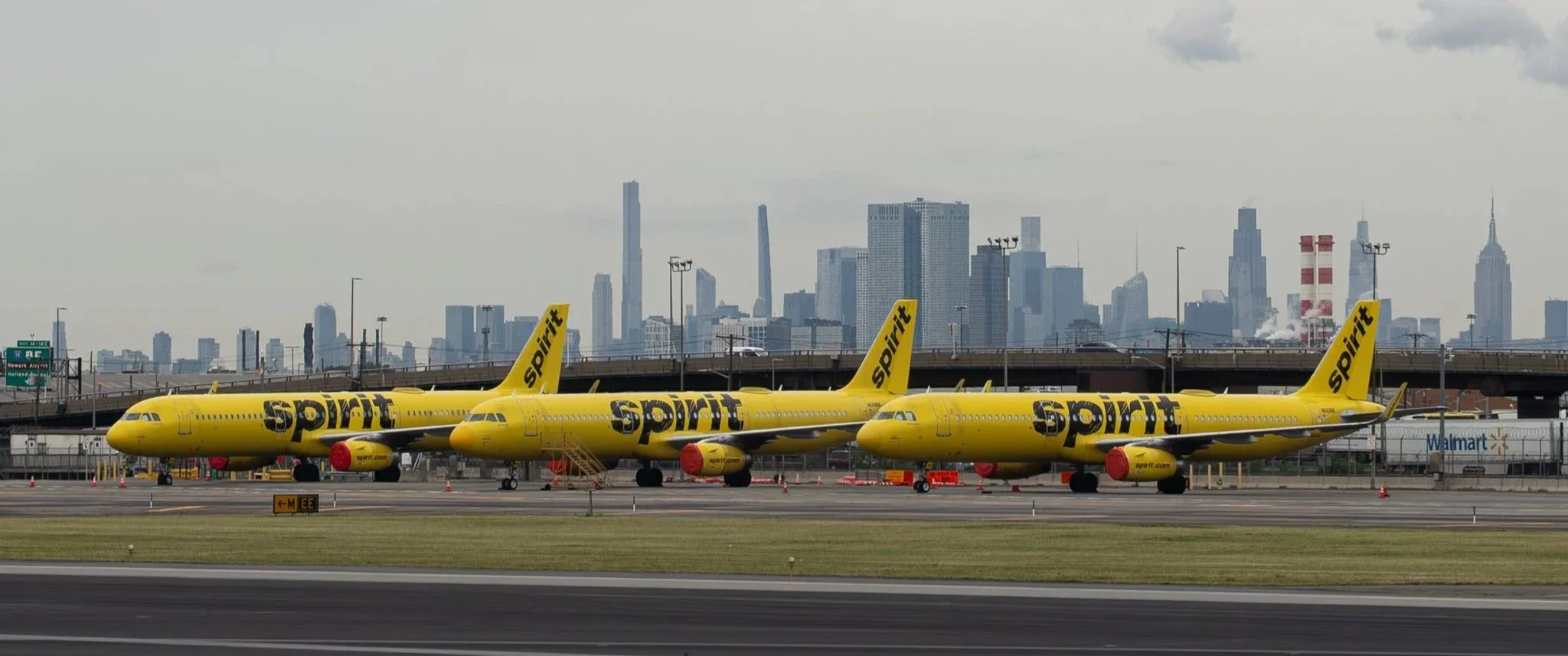 Three yellow Spirit Airlines airplanes parked on the tarmac with a city skyline in the background, including tall skyscrapers, and a bridge overhead.