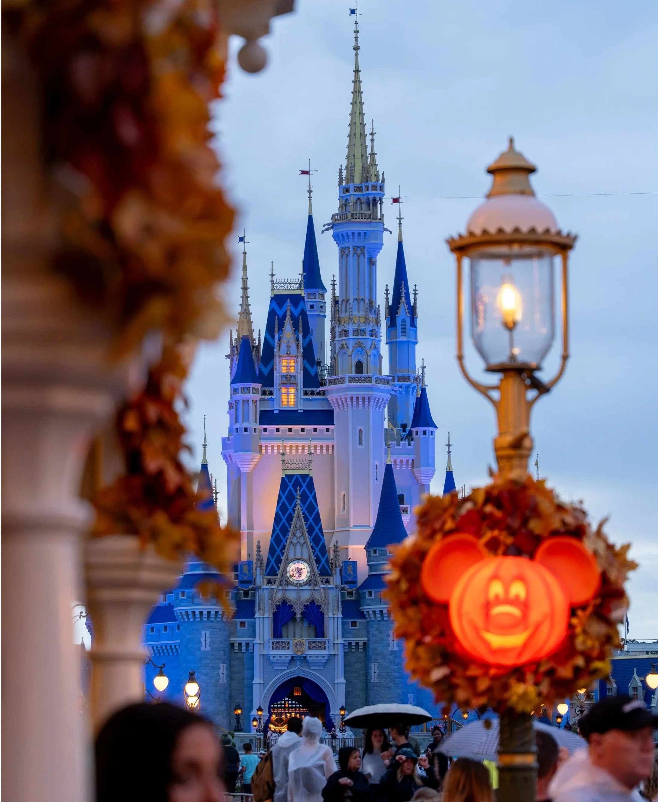 Disney castle illuminated at dusk viewed through Halloween-themed decorations, with visitors in rain gear walking underneath.