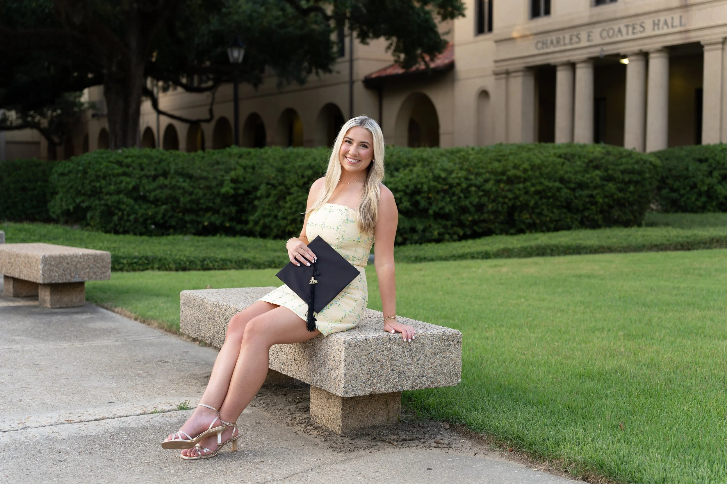 A young woman with long blonde hair, wearing a pale yellow dress, white high-heeled sandals, and holding a black graduation cap, sitting on a stone bench outdoors with green bushes and a large building labeled Charles E. Coates Hall in the background, smiling at the camera.