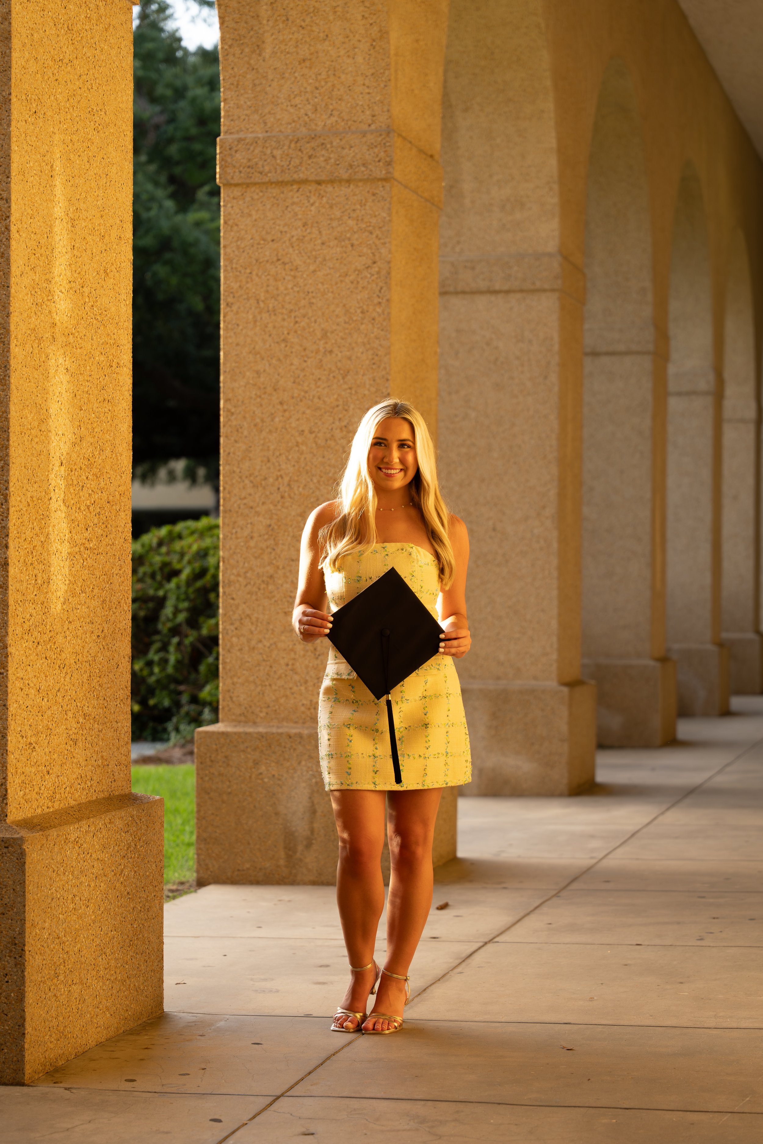 A young woman in a light yellow dress with a black graduation cap holding a diploma stands outside near a series of beige stone columns on a sunny day.