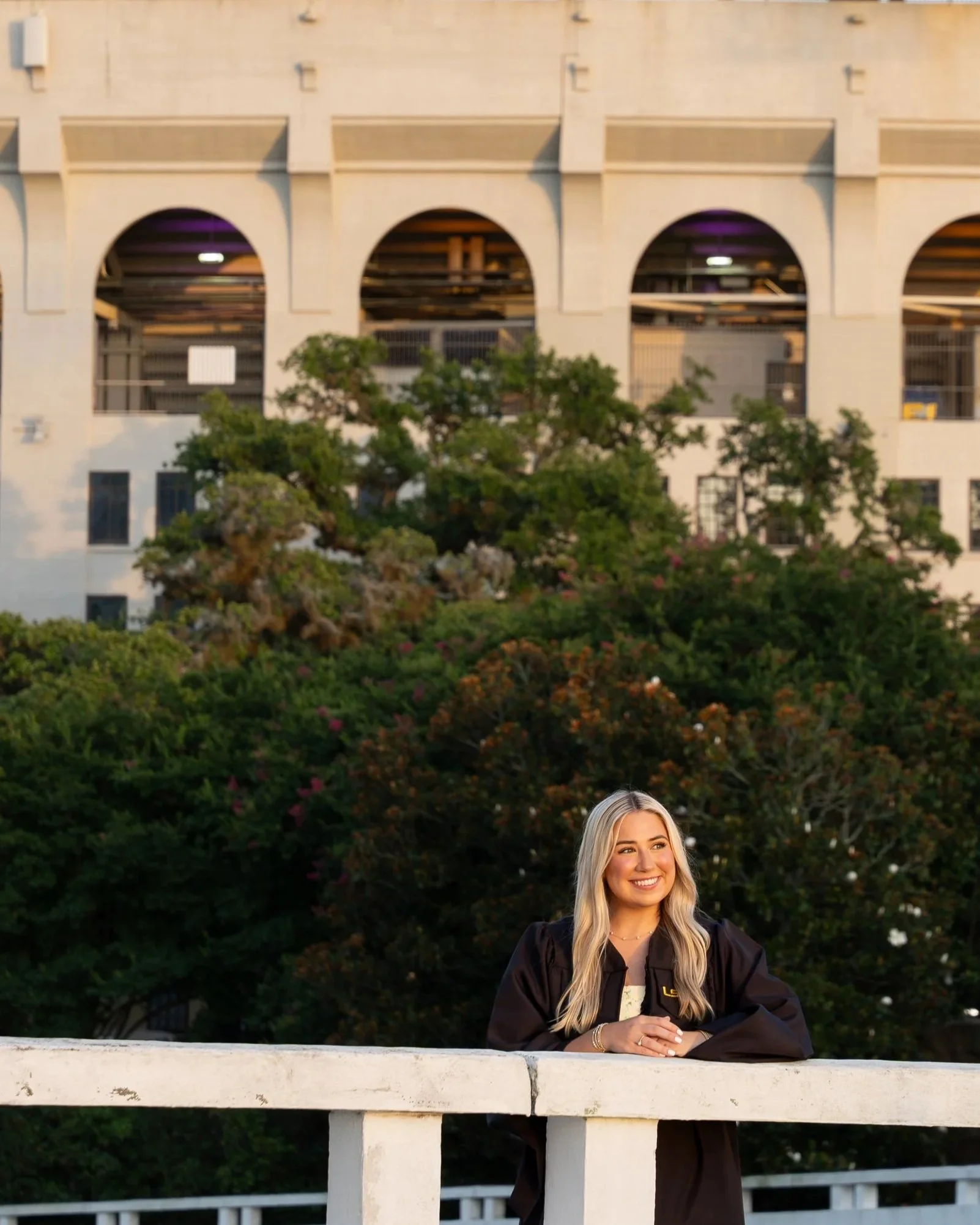 A young woman with long blonde hair smiling, wearing a black graduation gown, sitting outdoors on a white concrete ledge with greenery and trees in the background.