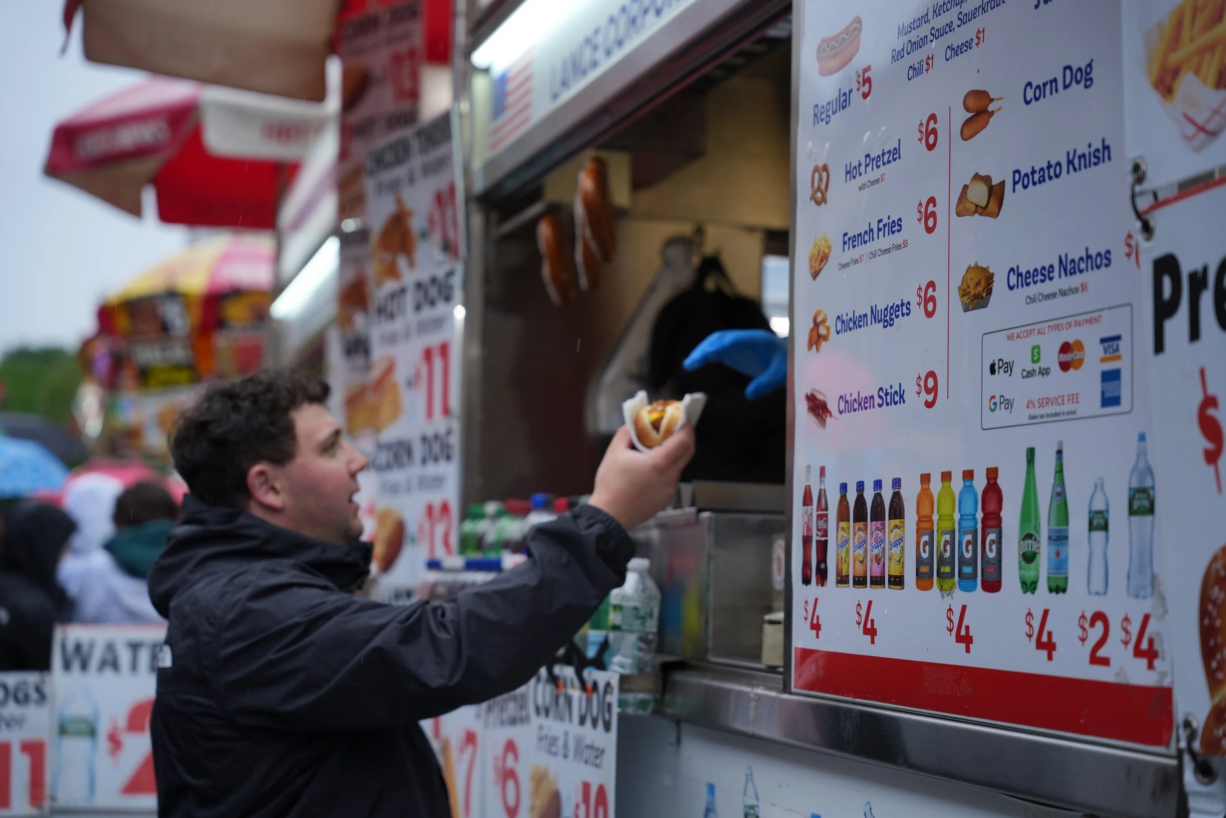 A young man with curly hair wearing a black jacket is holding a hot dog at a food stand. The stand has a large menu with food and drink items, prices, and images, and is busy with several people.