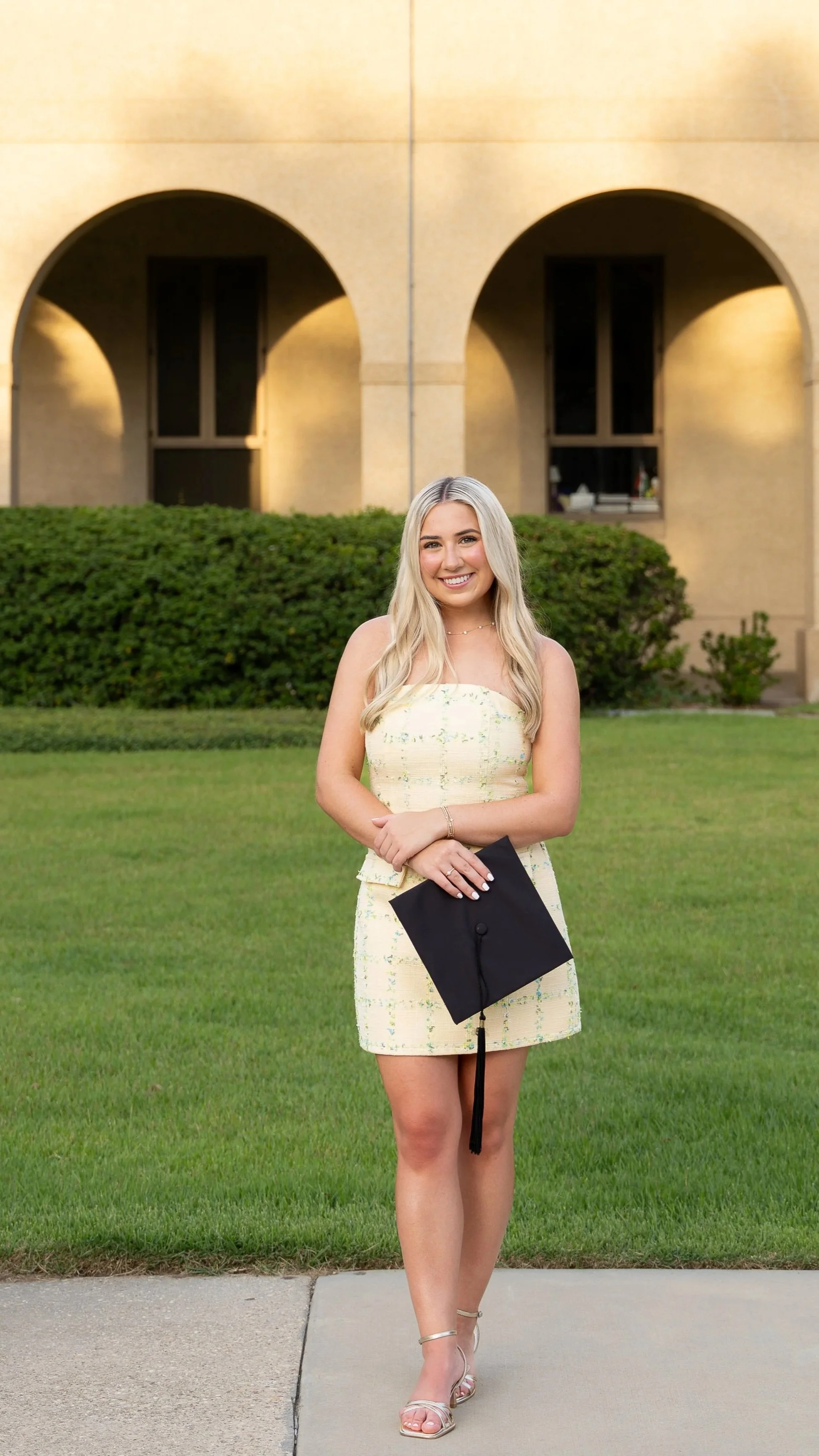 A young woman with long blonde hair in a yellow dress holding a graduation cap, standing outdoors on a sidewalk with green grass and bushes, and a building with arched windows in the background during daytime.