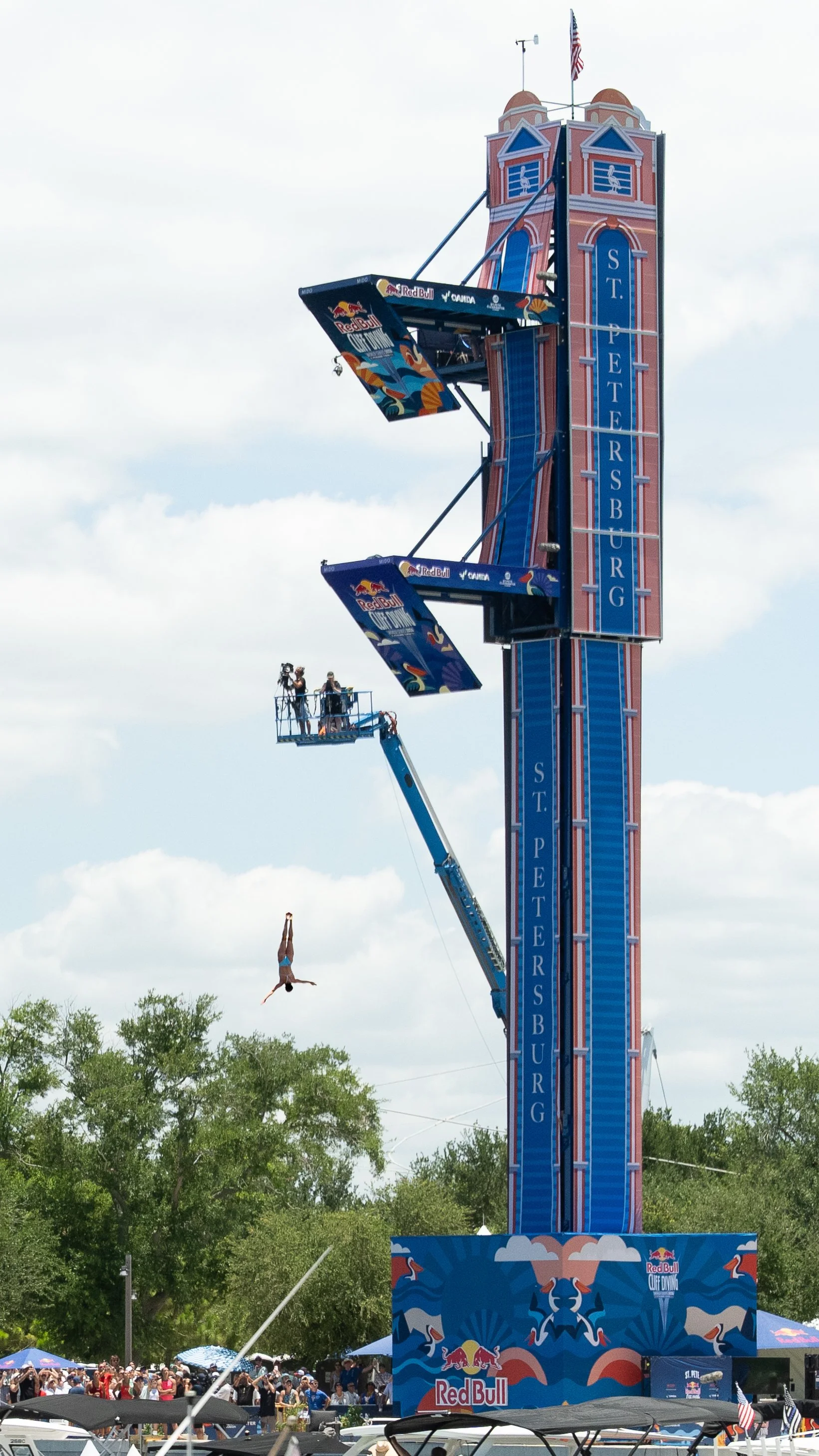 A person is mid-air, diving headfirst from a tall amusement park ride structure labeled 'St. Peter's BURN' at a Red Bull cliff diving event. There is a large crowd below watching the event, with trees and a cloudy sky in the background.