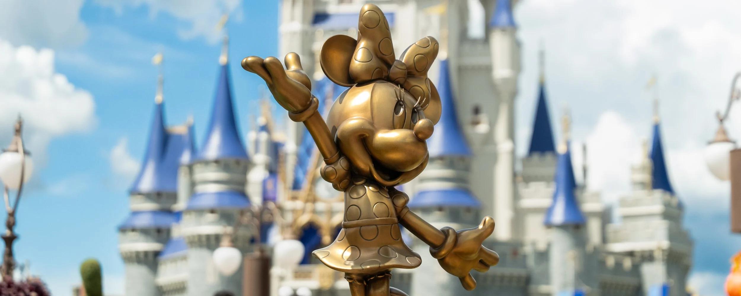 Golden statue of Minnie Mouse with a bow on her head, waving, in front of Sleeping Beauty Castle at Disneyland.