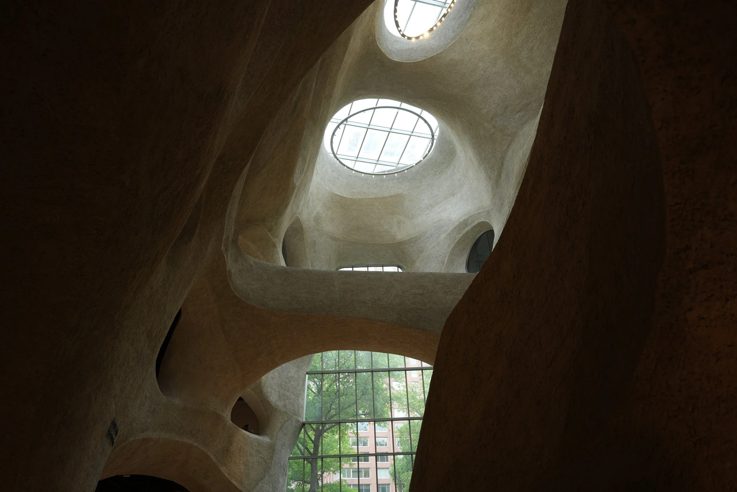 Looking up from inside a modern building with curved, beige walls and large oval skylights with glass and metal framework letting in natural light.