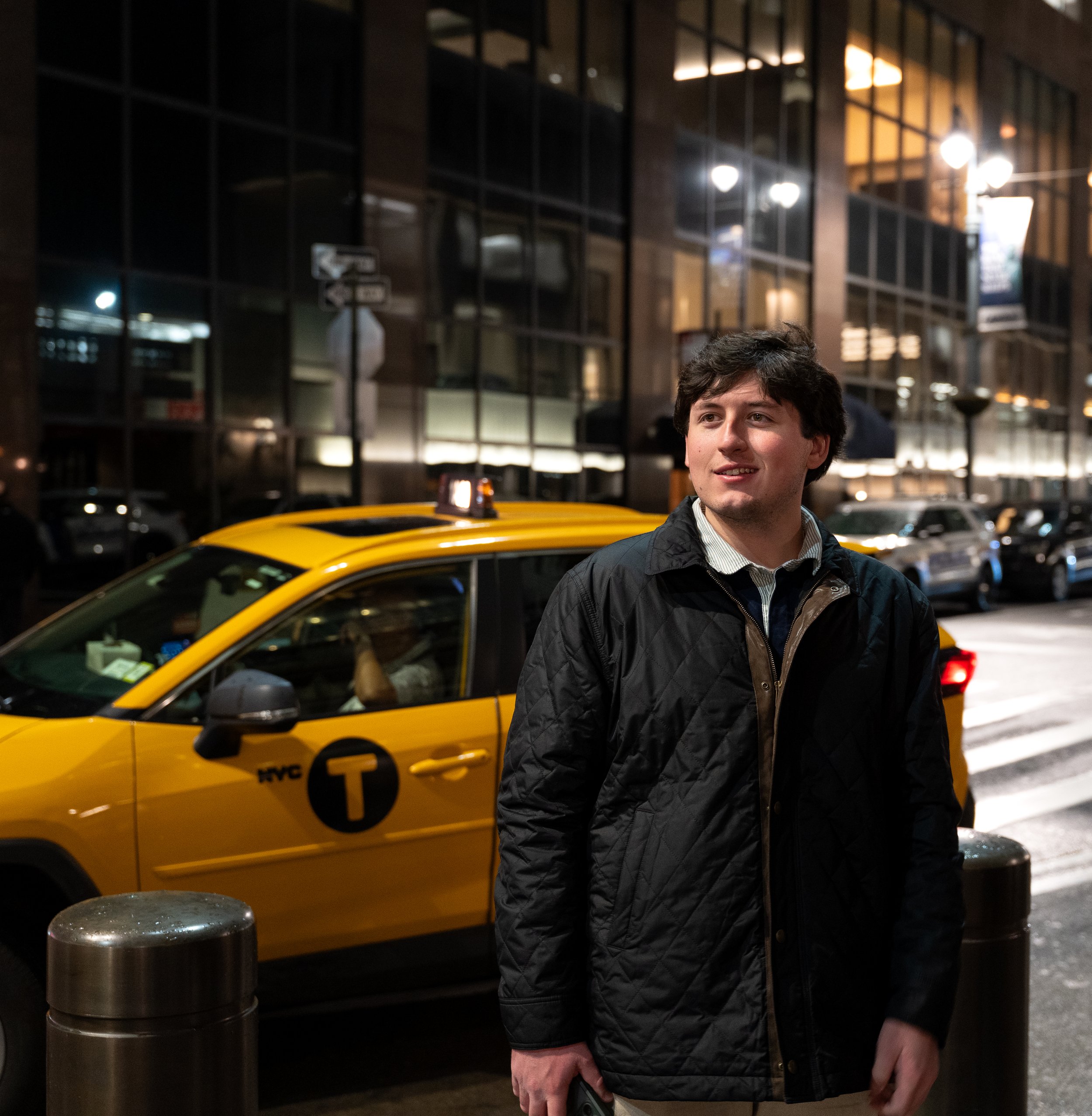A young man with curly dark hair smiling outdoors at night, standing near a yellow NYC taxi, in front of a modern glass building with bright lights.