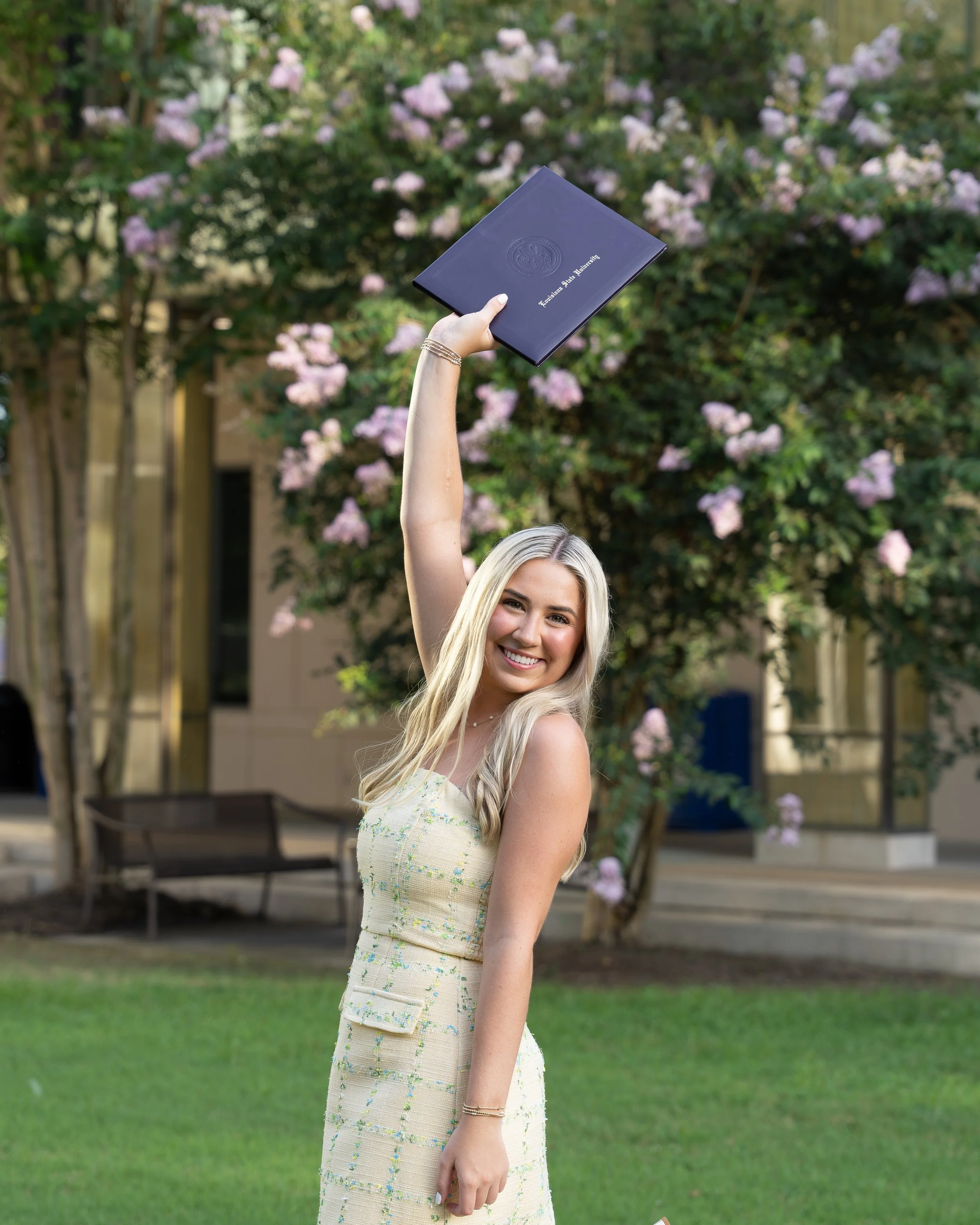 A young woman with blonde hair smiling, holding a diploma from Louisiana State University, outdoors in front of trees with pink flowers.