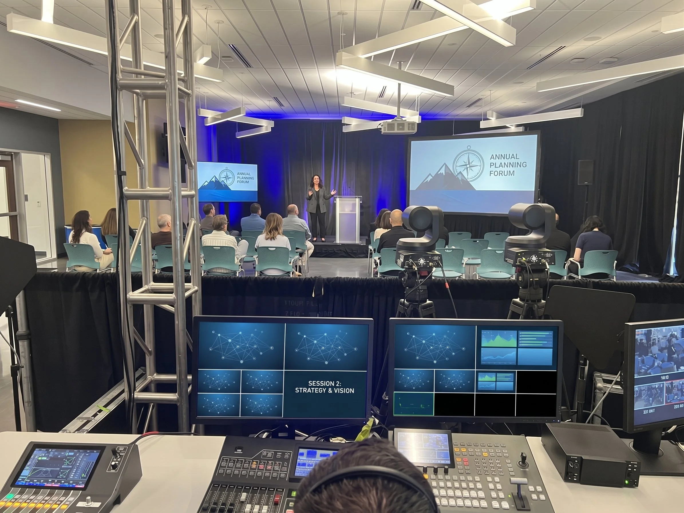 A conference room with a stage and large screens, showing a presentation titled "Annual Planning Forum." There are people seated facing the stage, and the room is equipped with technical equipment and lighting fixtures.