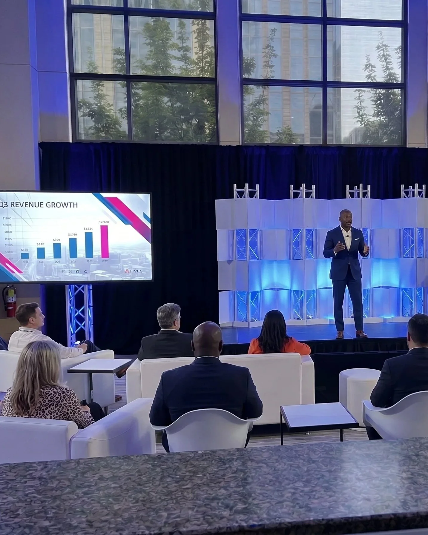 A speaker in a navy blue suit giving a presentation on stage at a conference. A large screen beside him displays a bar graph labeled 'Q3 Revenue Growth' with financial figures. The audience sits in white chairs, attentively listening. The room has large windows showing buildings and trees outside.