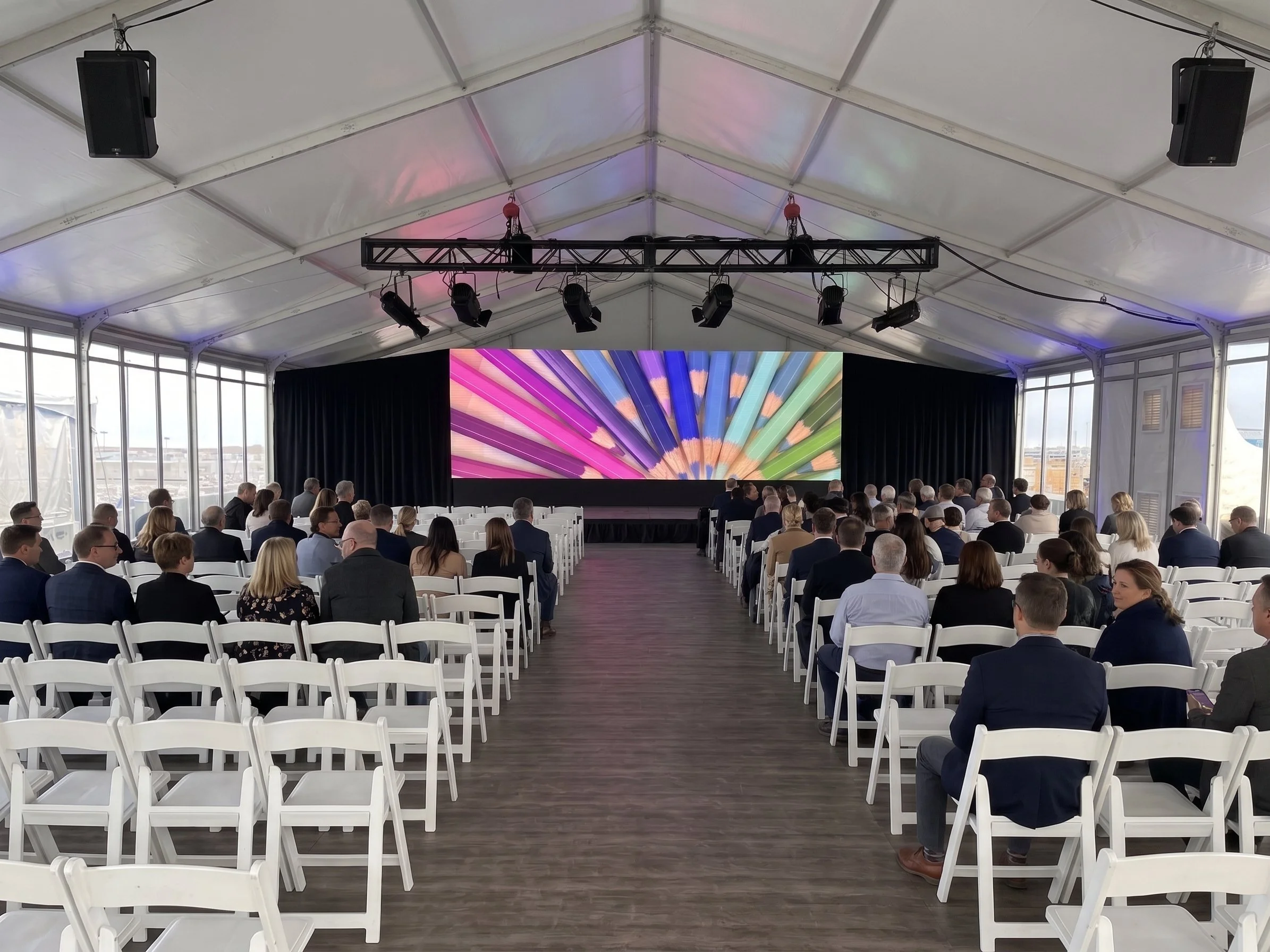 A large conference or event tent filled with people seated in white chairs facing a stage with a colorful digital display. The tent has windows on the sides, and lighting and sound equipment are installed on a truss hanging from the ceiling.