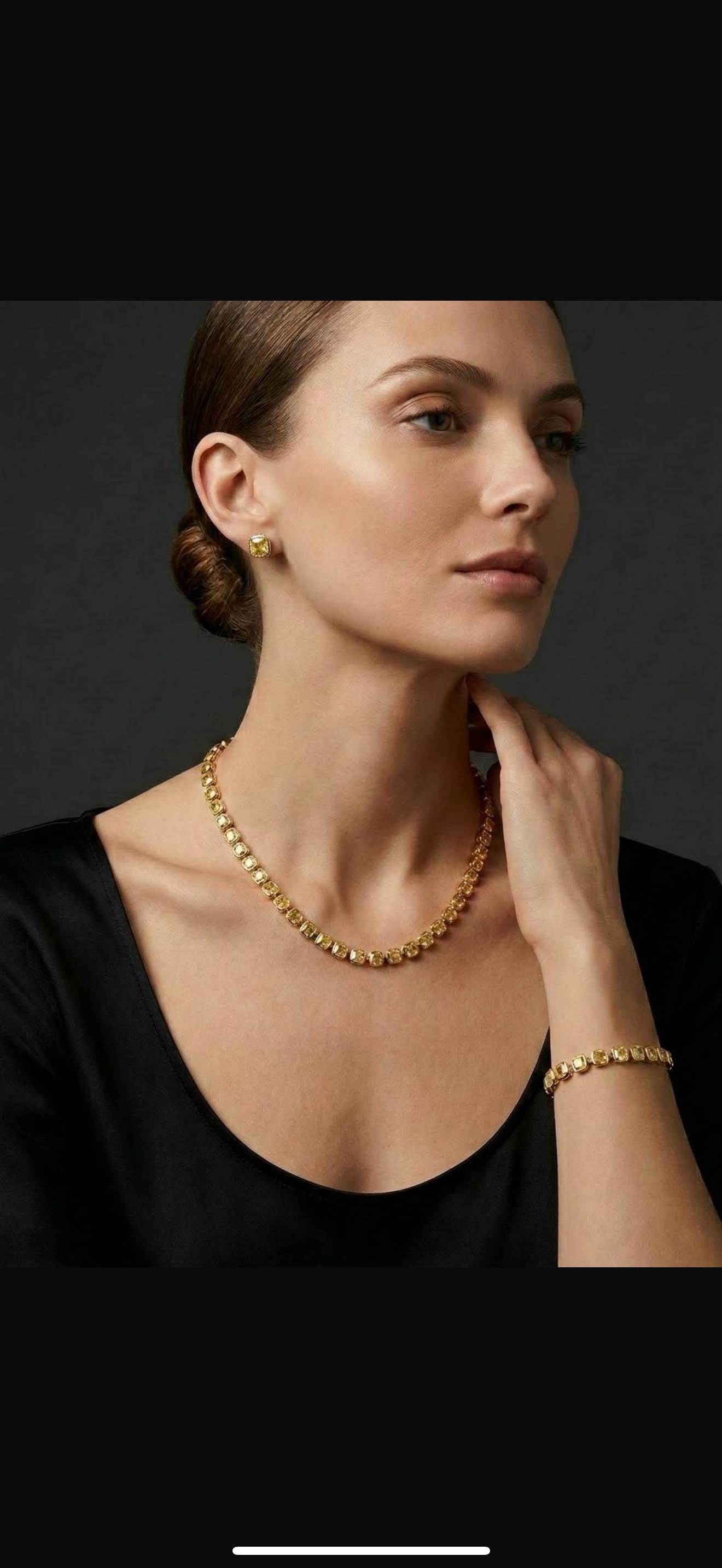 Woman wearing gold jewelry, including earrings, a necklace, a bracelet, and a ring, against a dark background.