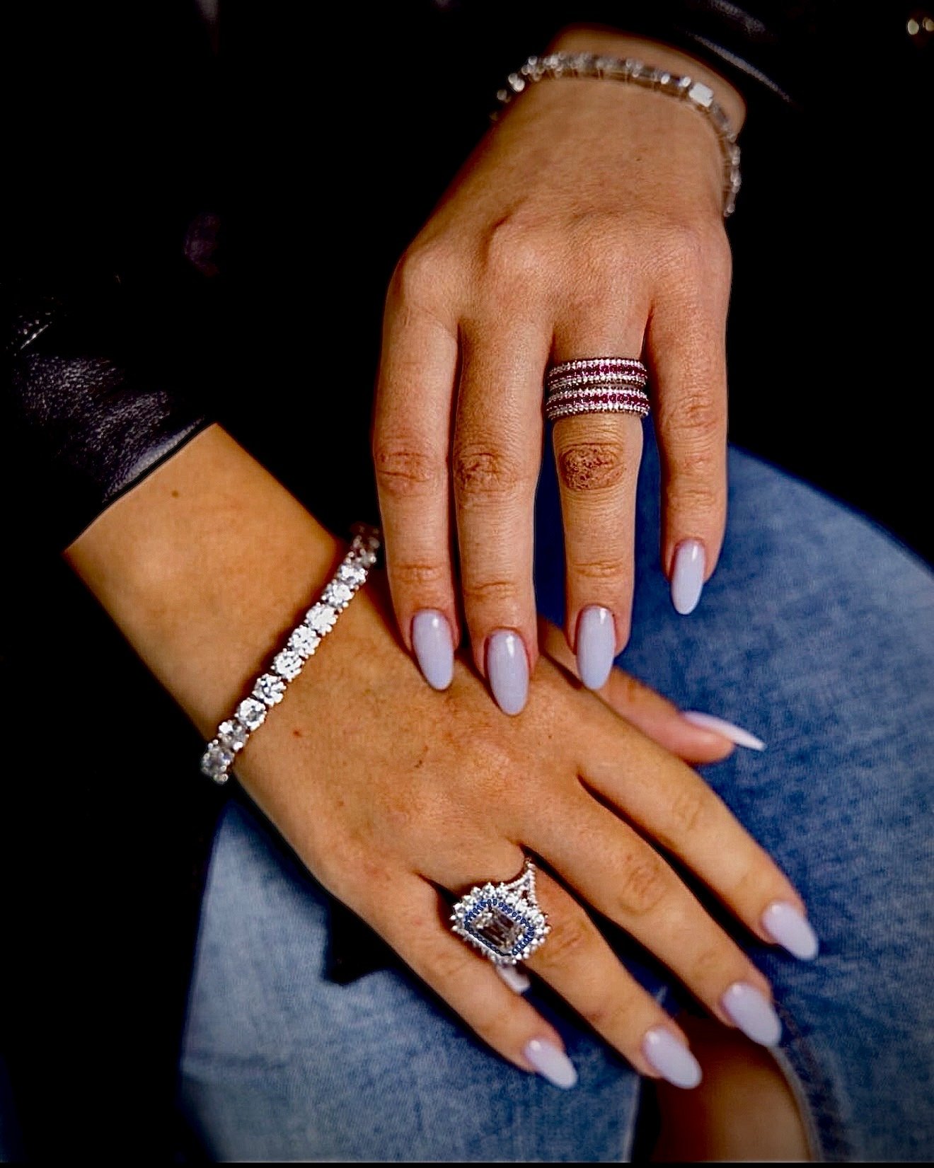 Close-up of a woman's hands resting on her lap, adorned with jewelry including rings, bracelets, and a watch, with manicured nails painted in a light shade.