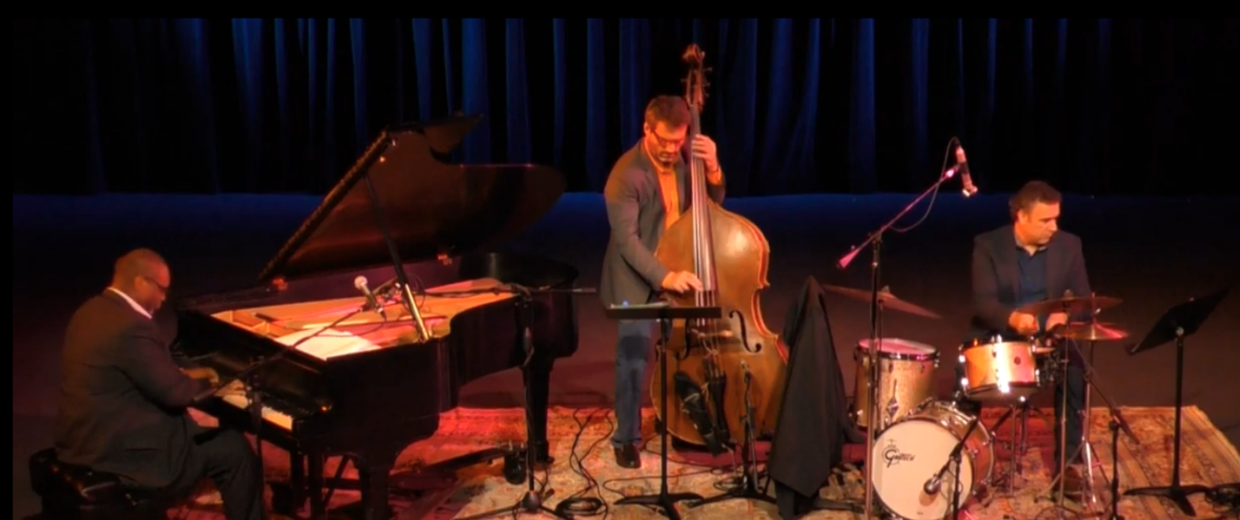 Trio Nexus on stage performing on their respective instruments.  From left to right, Reggie Thomas on grand piano, Dennis Carroll on upright bass, and George Fludas on drumset.