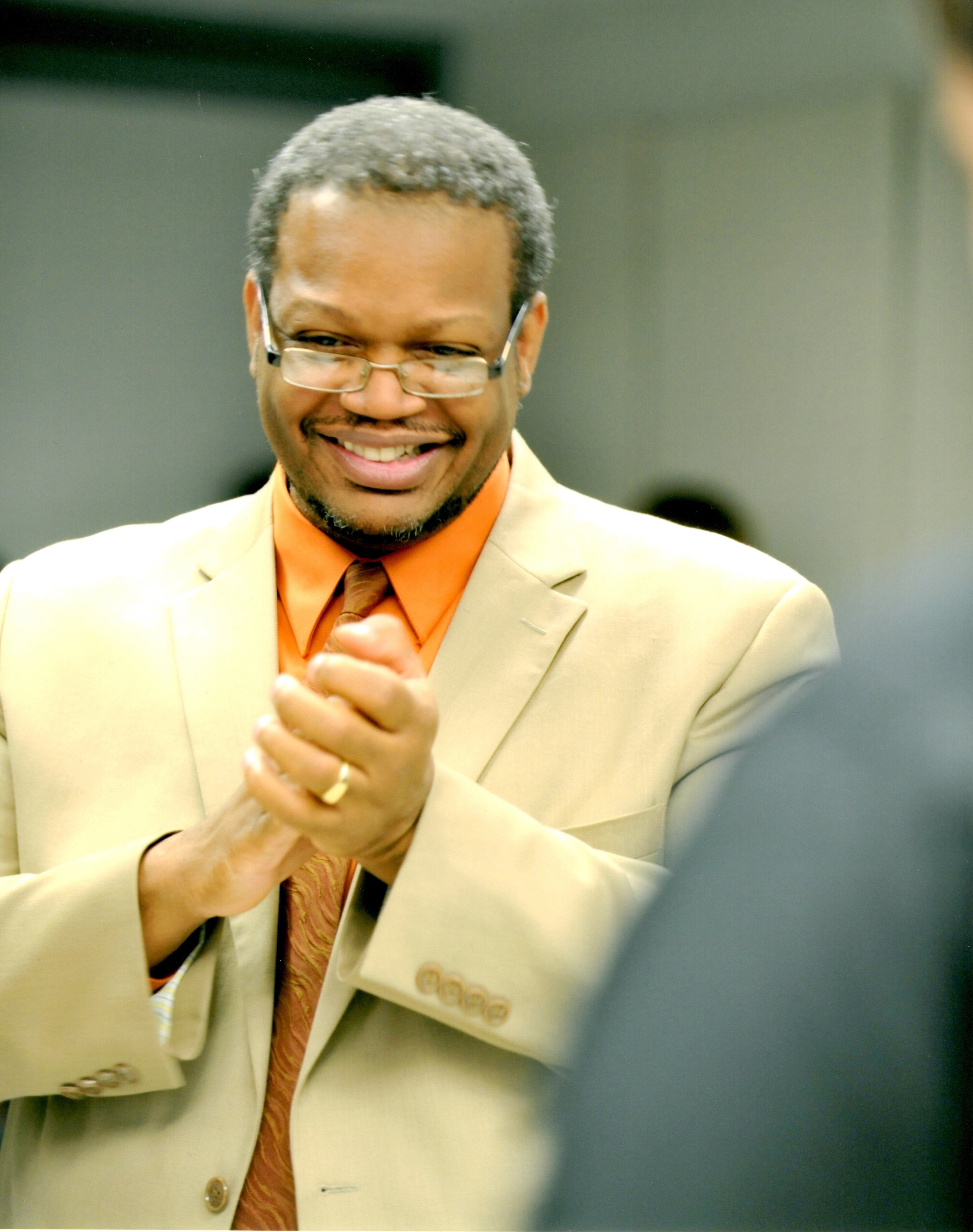 Reggie Thomas in a tan suit smiling with his hands together.