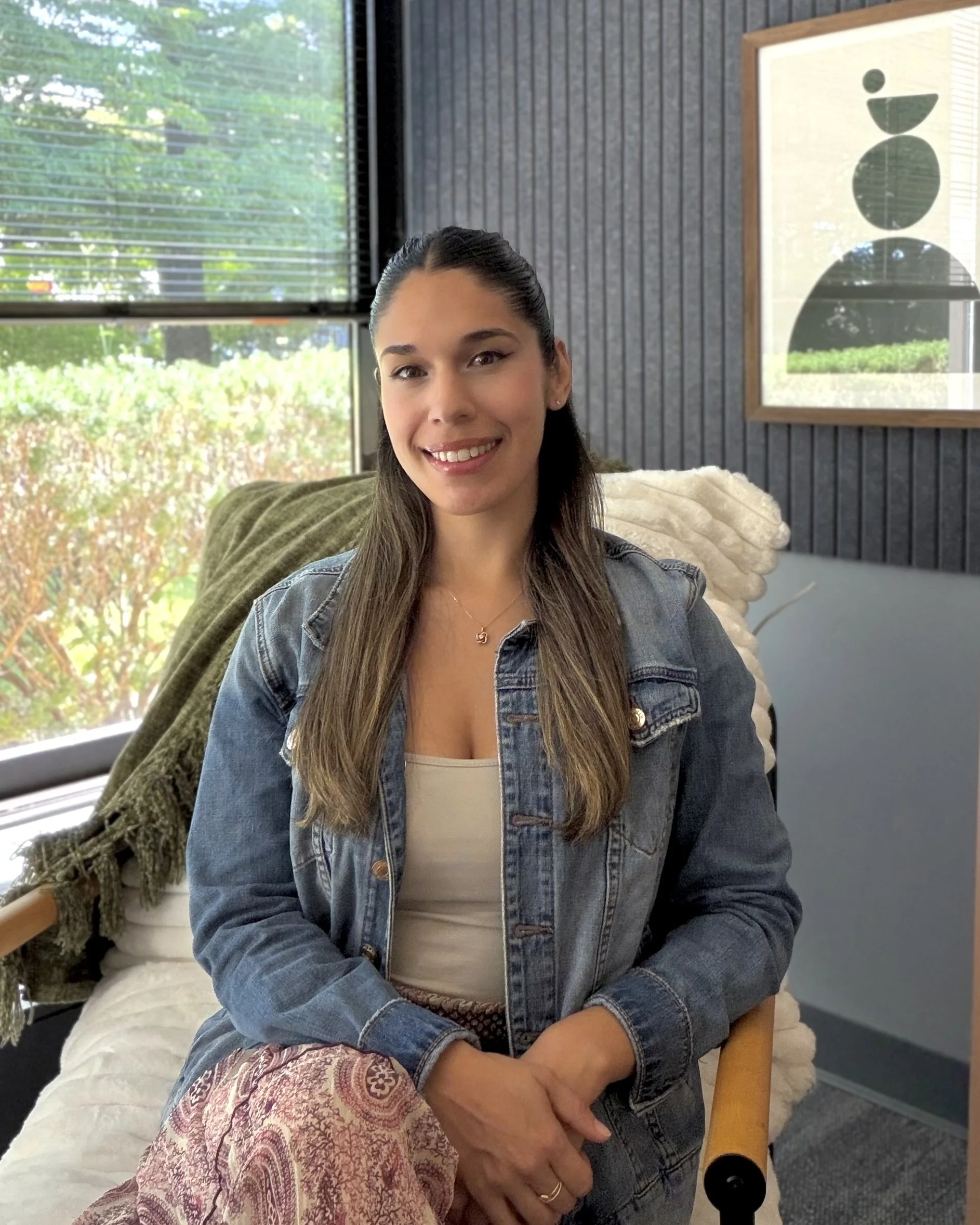 A young woman with long brown hair, wearing a denim jacket and a beige top, smiling while sitting in front of a window with blinds. There's outdoor greenery visible through the window and a framed abstract art piece with black shapes on a light background hanging on the wall behind her. She appears to be seated on a chair with a wooden armrest, and a patterned pillow is visible on her lap.
