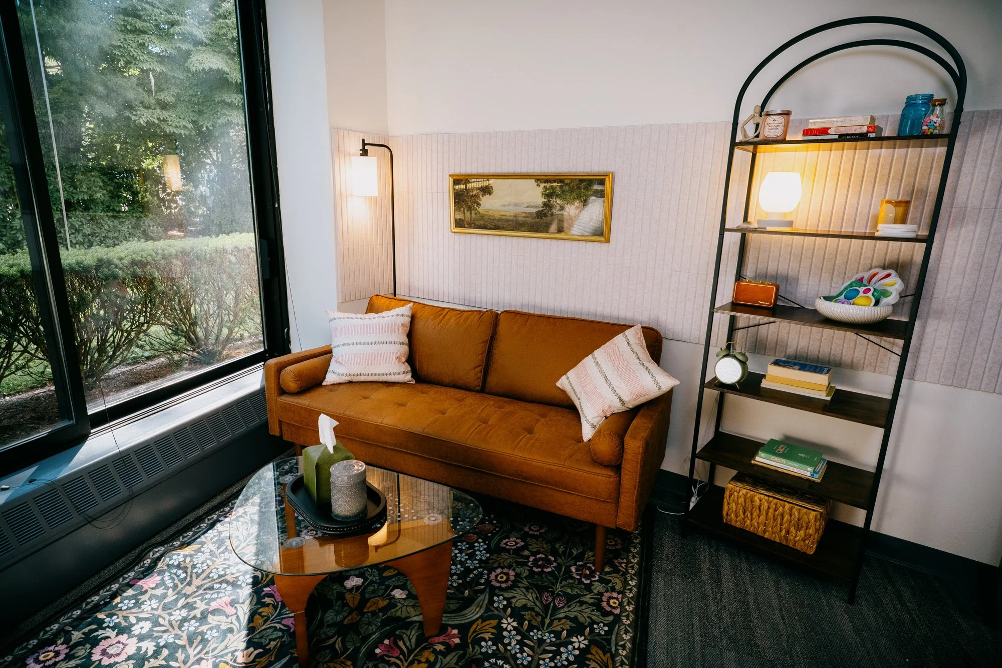 A cozy living room corner with a mustard brown sofa decorated with striped pillows, a glass coffee table with candles, a large window with a view of greenery, a tall black shelf with books and decorative items, a wall lamp, and a framed landscape painting on the wall.