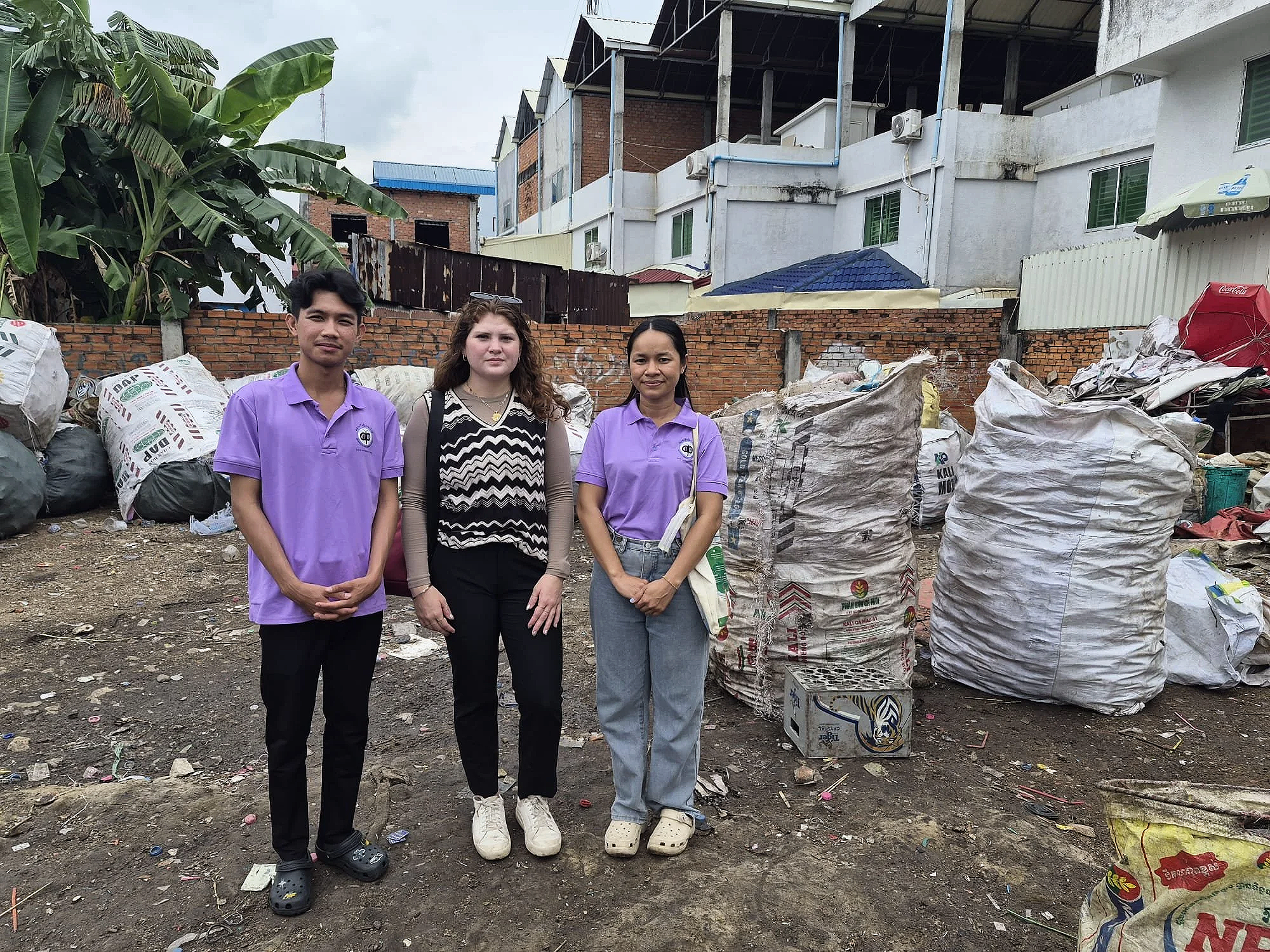 Three people standing outdoors in a trash-filled area with bags of waste behind them.