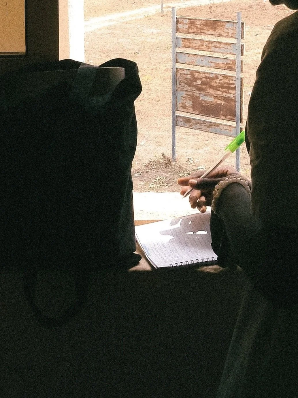 A person writing on a notepad with a pen, sitting at a desk near a window with an outside view of a dirt area and a wooden sign or structure.
