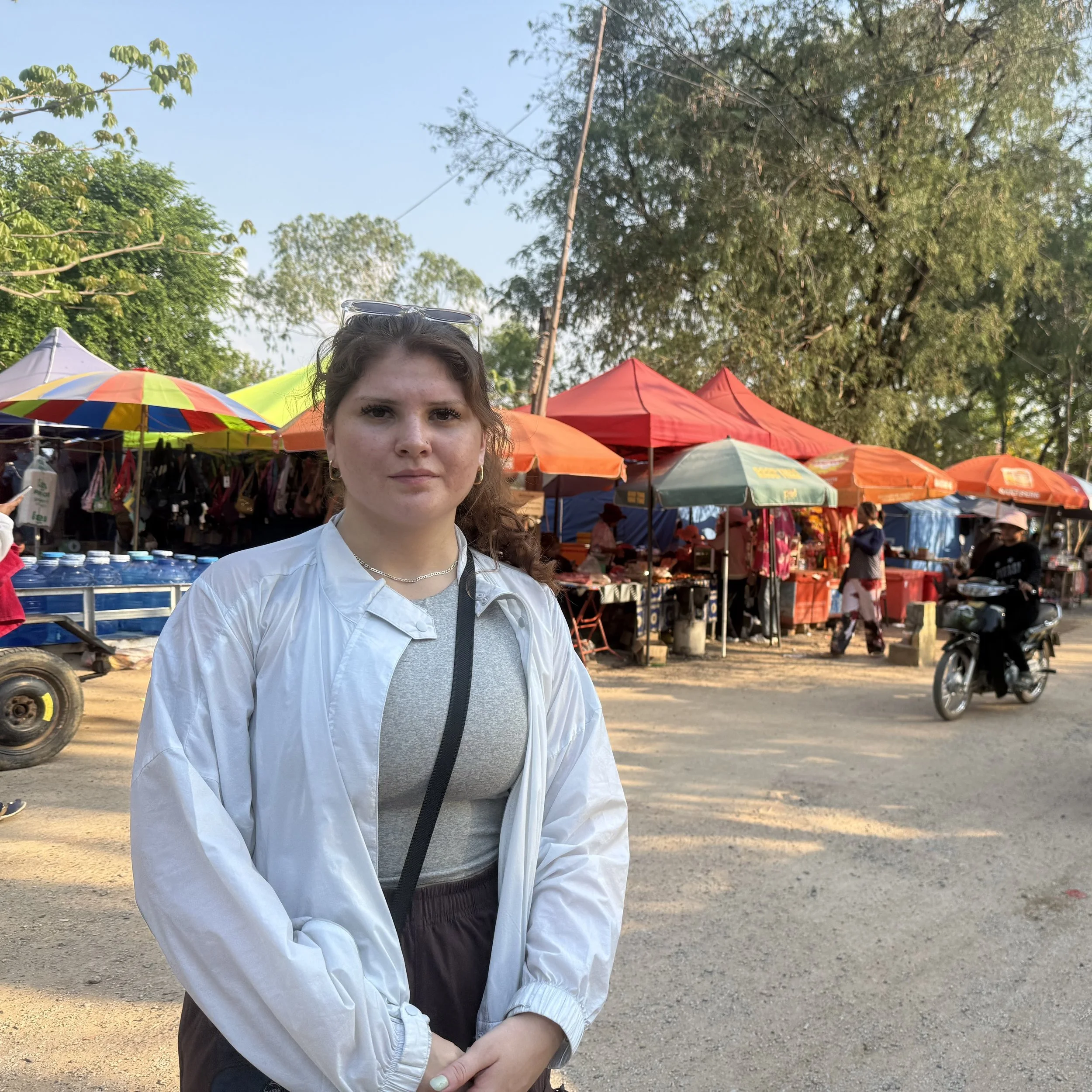 A woman with brown hair, wearing a light gray shirt, sunglasses on her head, and a white jacket, stands outdoors at a market with colorful umbrellas and trees in the background.