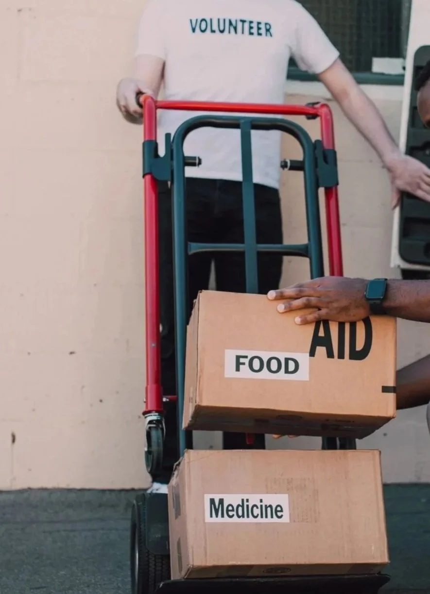 Person in a white t-shirt labeled 'VOLUNTEER' pushing a grocery cart with labeled boxes containing food, aid, and medicine.