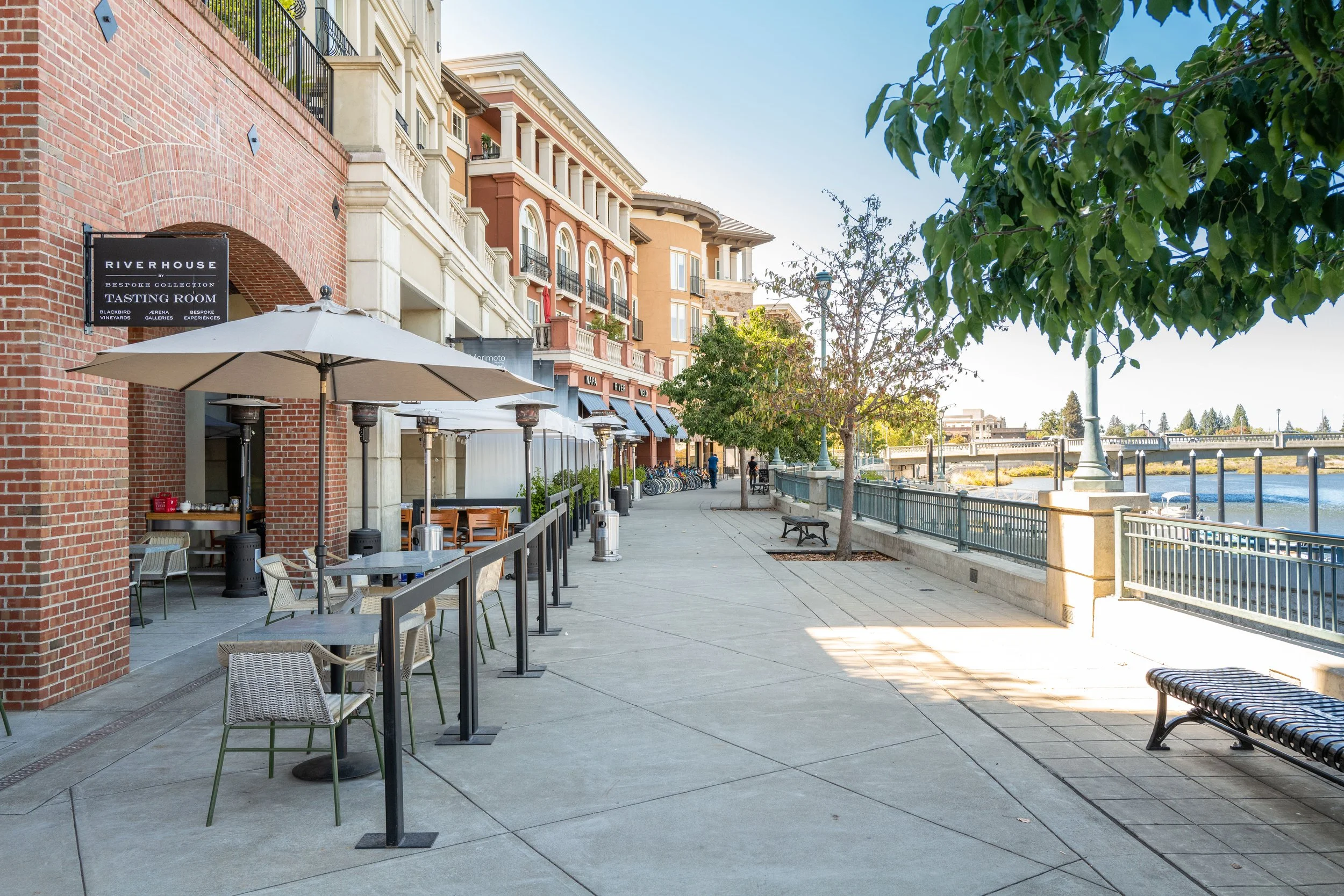 Empty outdoor sidewalk along a river with tables, chairs, umbrellas, trees, benches, and colorful buildings in the background on a sunny day.