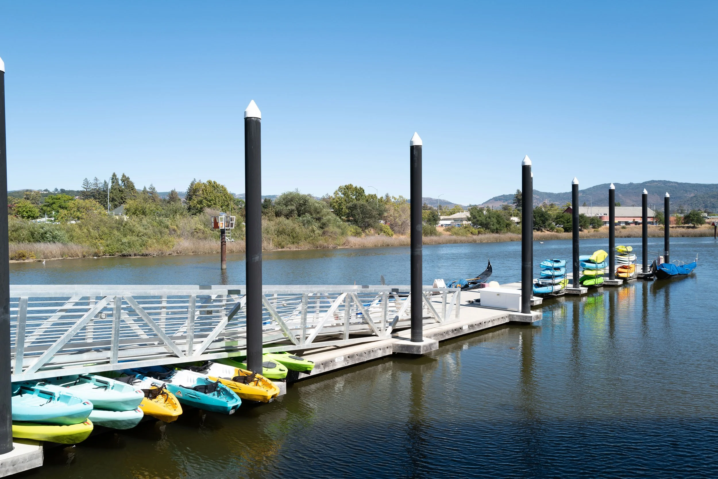 Dock with colorful kayaks and paddleboards on a calm river under a blue sky with trees and hills in the background.