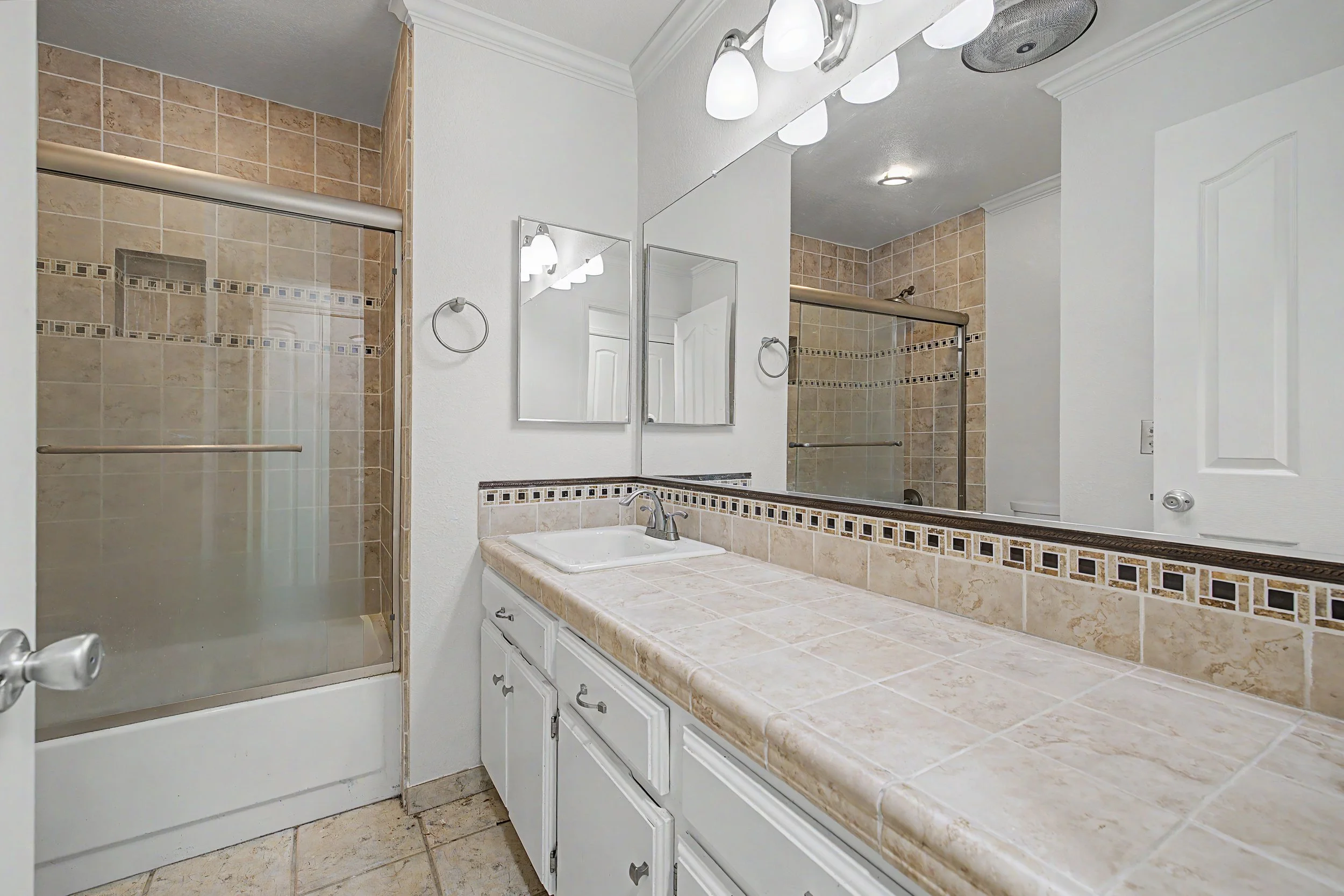 A bathroom with a shower area, countertop with a sink, large mirror, and tile decor, including a towel ring and shelves.