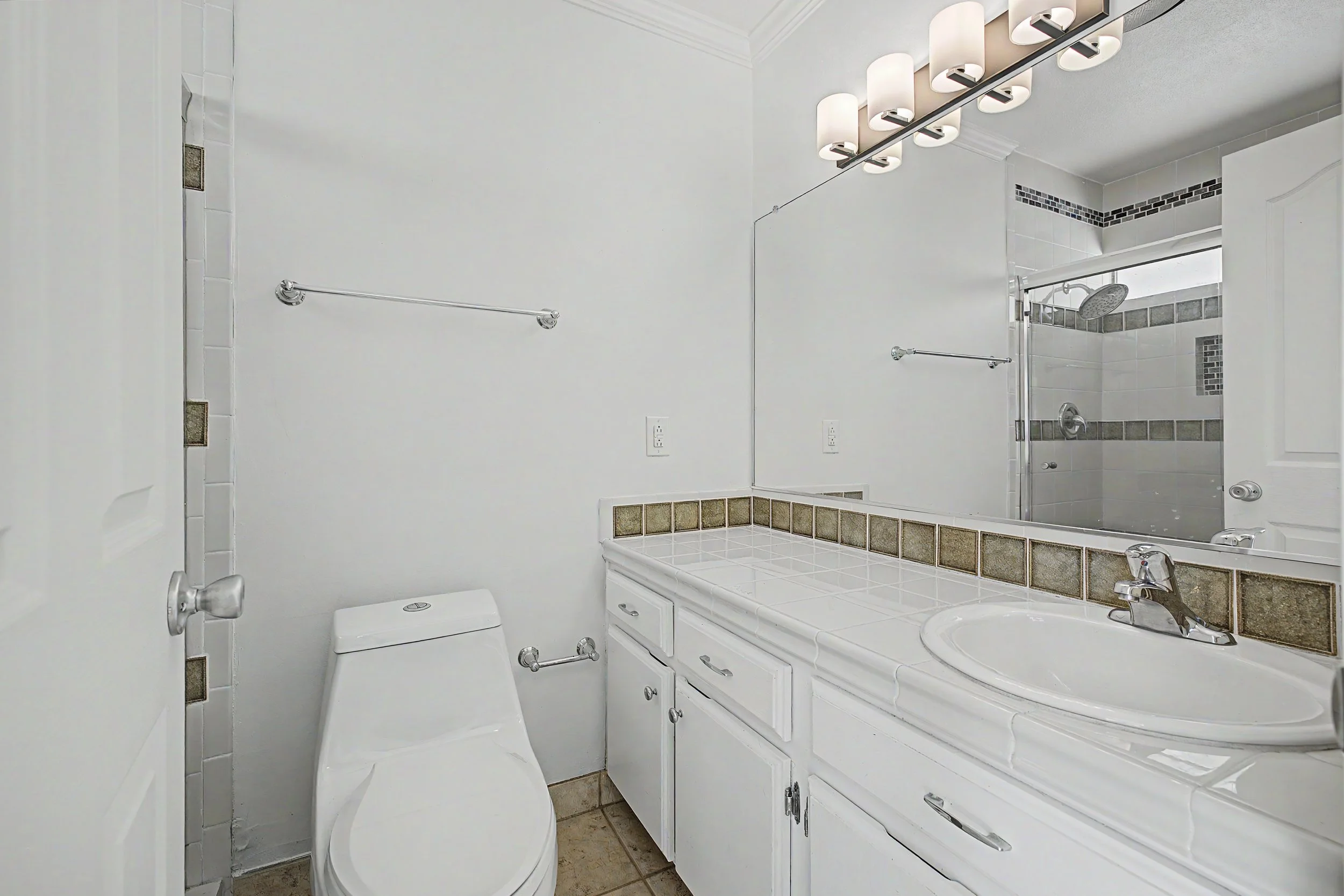 A small bathroom with a white toilet, a white vanity with a sink, and a mirror above the sink. There are white walls and a tiled shower area with decorative tile accents. The bathroom has a modern light fixture above the mirror.