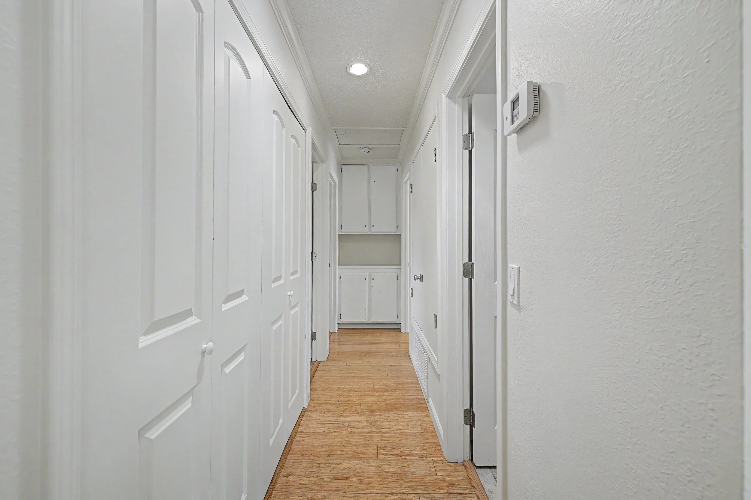 Empty hallway with white walls and white doors on the left side, wooden floor, small closet at the end, and textured ceiling with ceiling light and security system on the right wall.