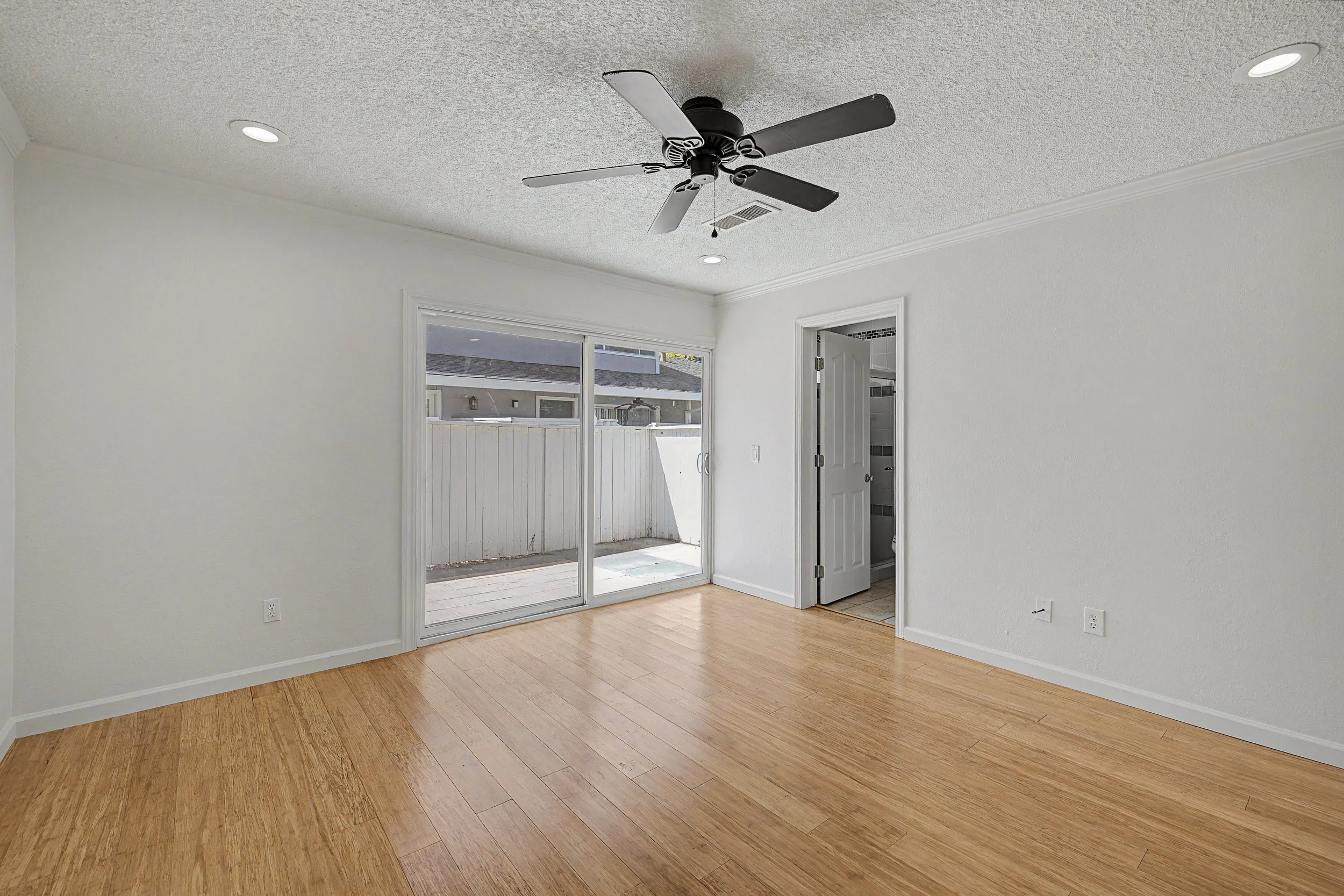 Empty room with hardwood floors, white walls, a ceiling fan, and a sliding glass door leading to a backyard.
