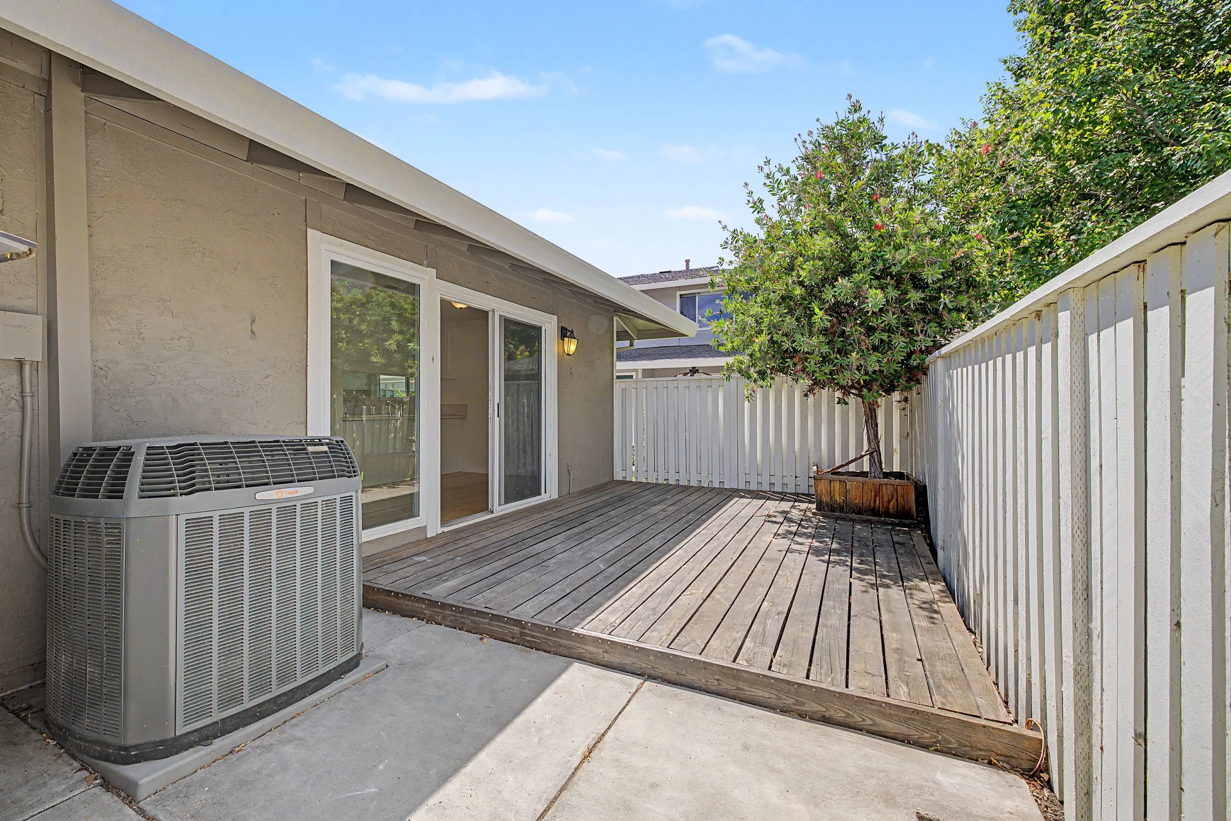 A backyard patio with a wooden deck, a potted tree, a white fence, and an air conditioning unit. The house has a sliding glass door and an exterior light fixture. The sky is blue with a few clouds.