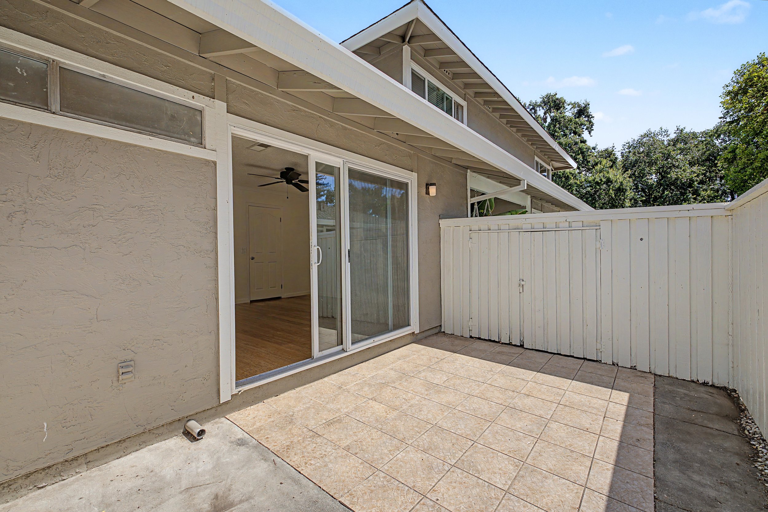 Exterior view of a house showing a sliding glass door leading to a patio with beige tiles, a white wooden fence, and part of the house's beige stucco wall. The house has a two-story structure with a small window near the roof and is surrounded by gre