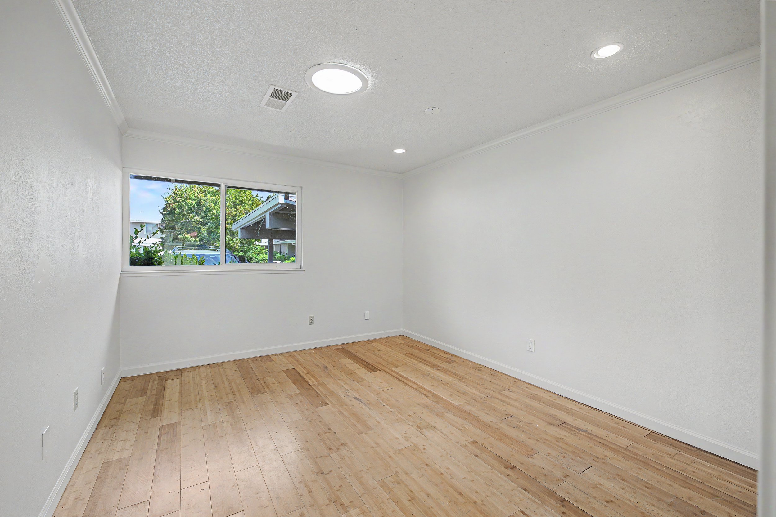 Empty room with hardwood floors, white walls, and a window showing trees and a neighboring building. Ceiling has recessed lighting and air vent.